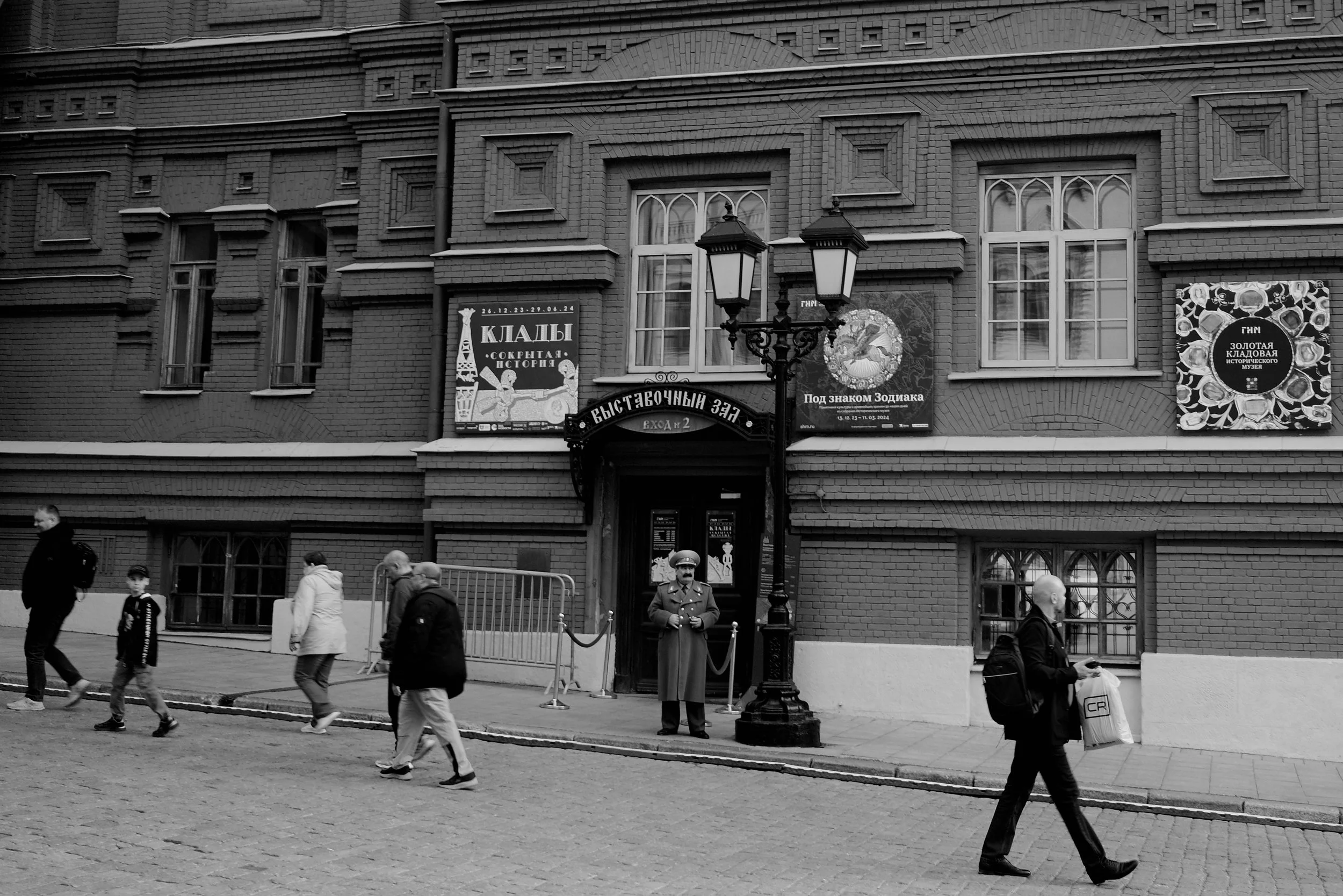 Black and white photo of a brick building with several posters and windows. Pedestrians walking on the cobblestone street, including a person with a backpack and a security guard standing near the entrance.