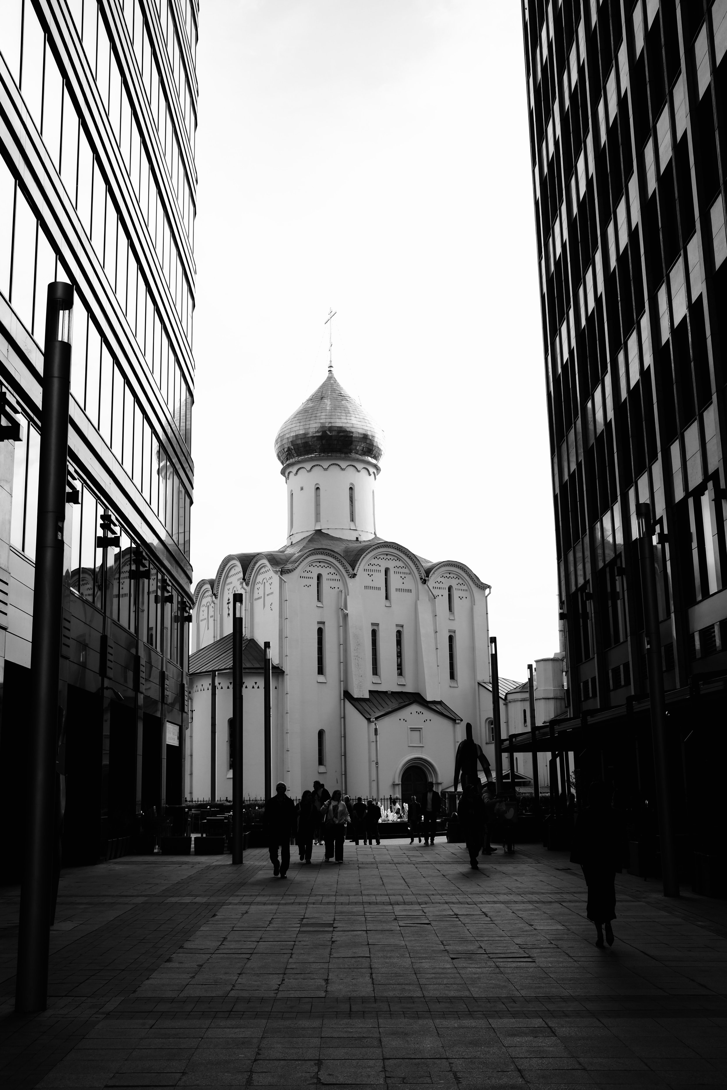 Black and white photo of a church with a domed roof, flanked by modern glass buildings, with people walking in the foreground.