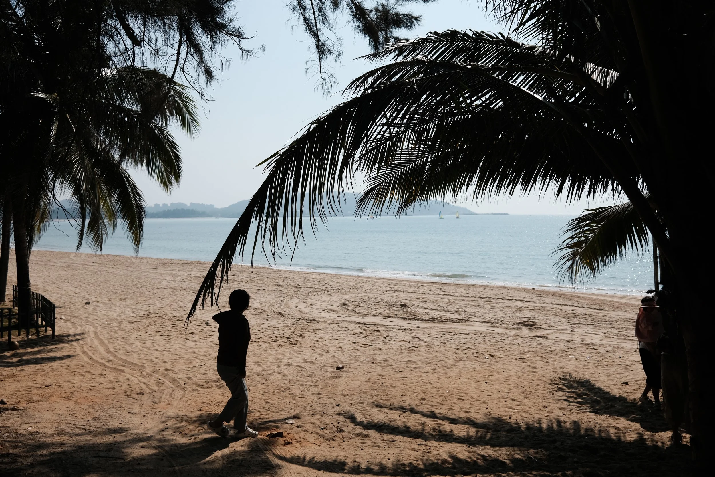 Silhouette of a person walking on a sandy beach shaded by palm trees, with water and distant sailboats in the background.