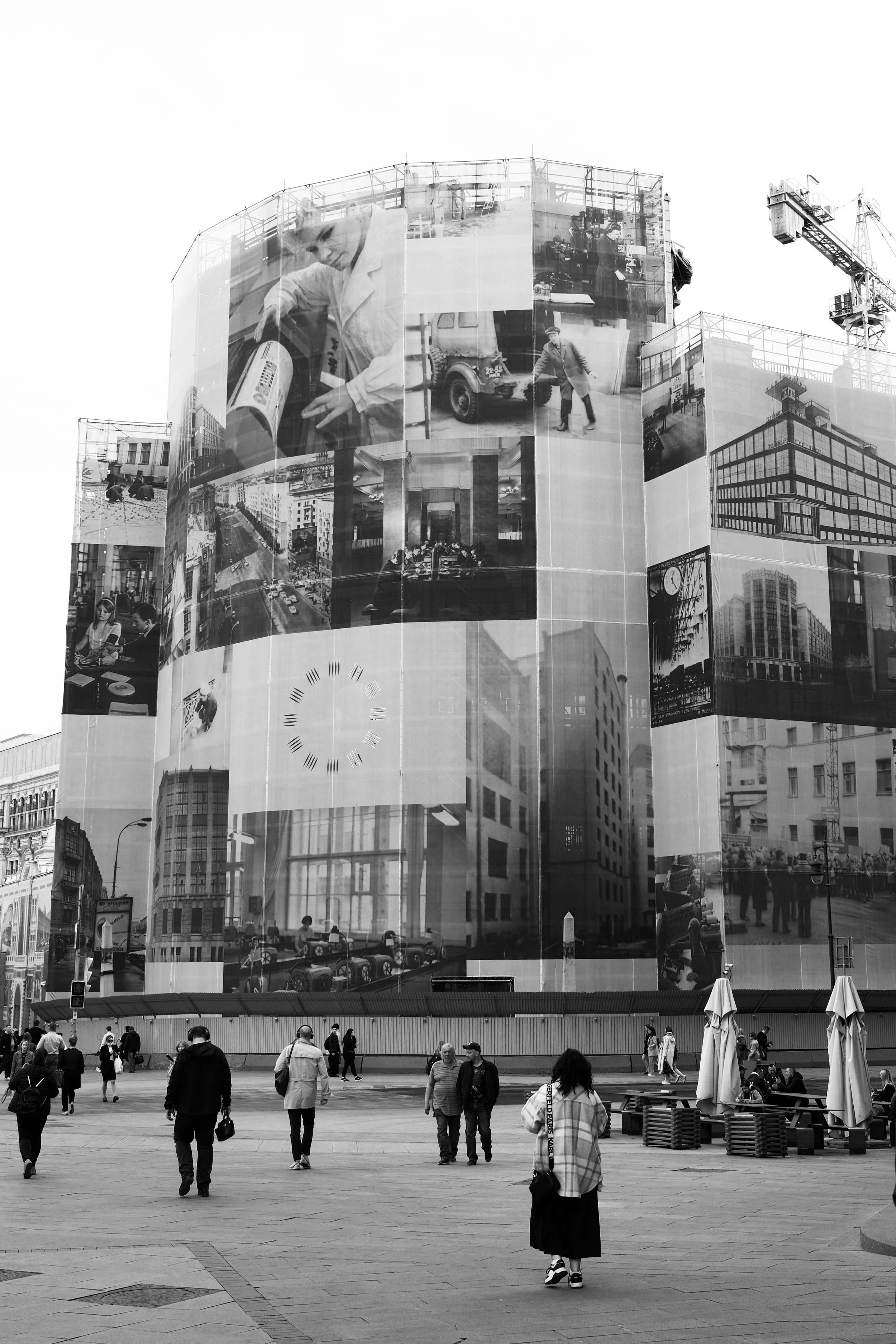 A black and white photo of a public square with people walking. A tall building in the background is covered with large banners displaying various images and a clock face. There are outdoor seating areas with umbrellas.