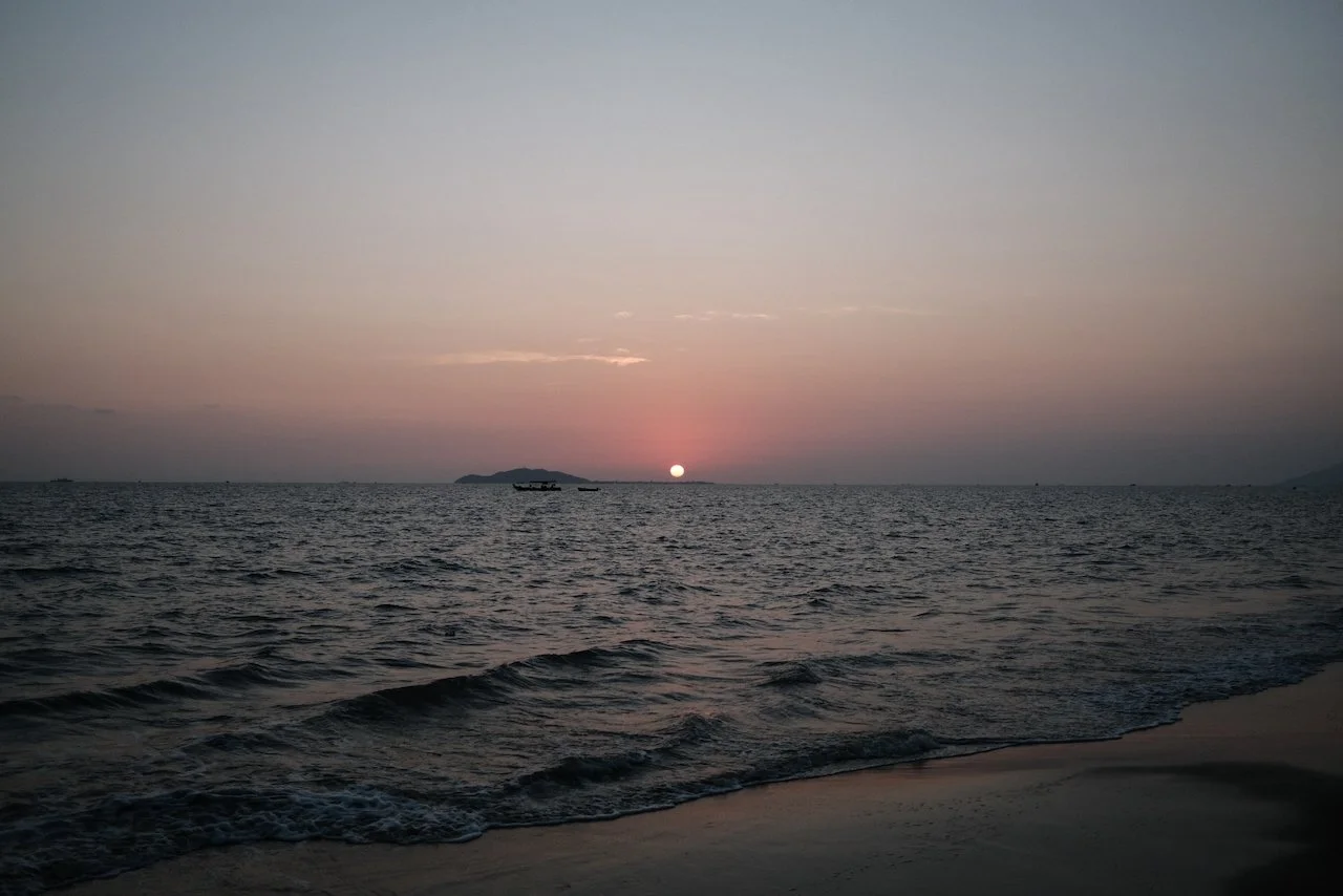Sunset over the ocean with small waves and a few boats in the distance.