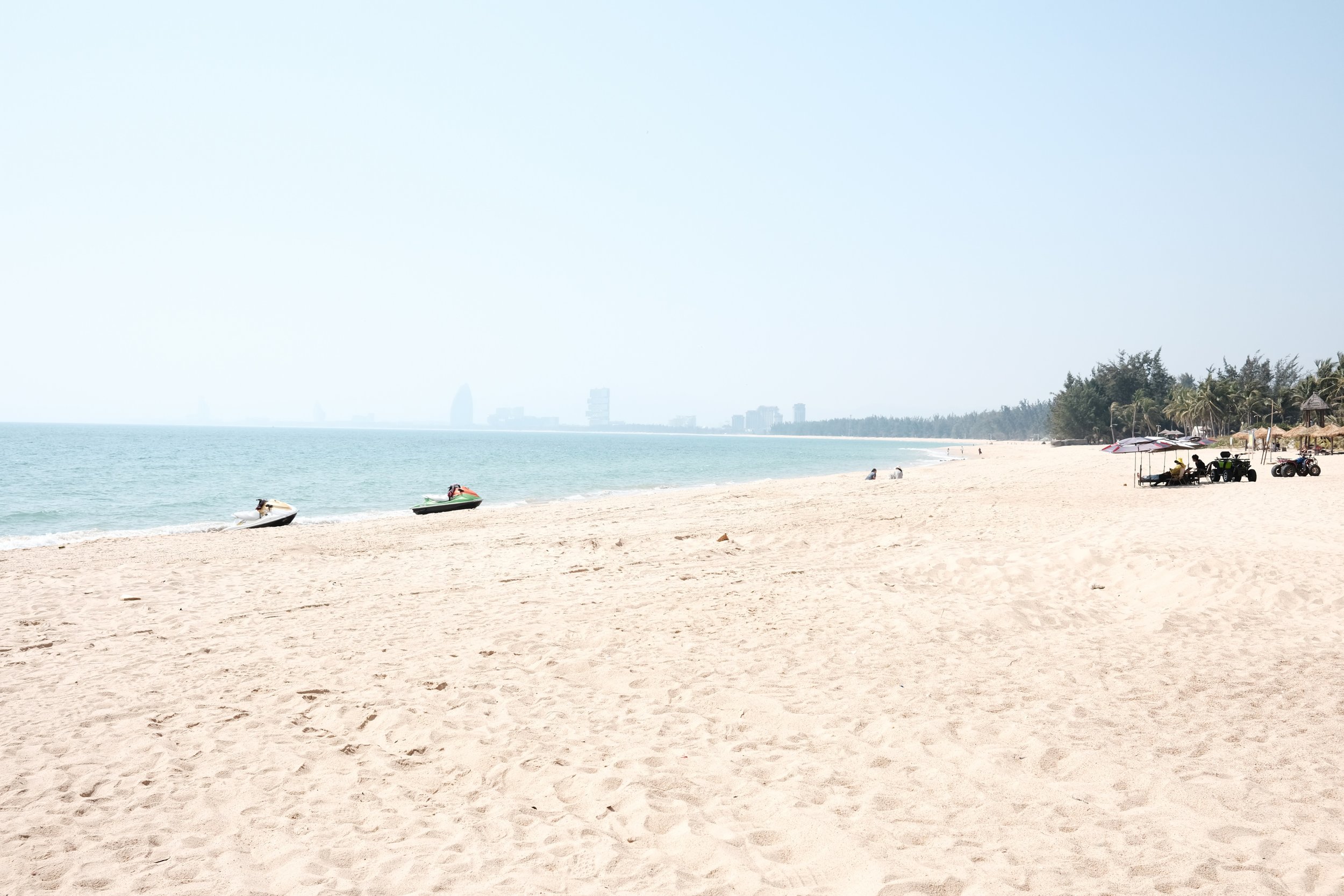 A sandy beach with a few people, colorful umbrellas, umbrellas, and trees, with buildings visible in the distance across the ocean.