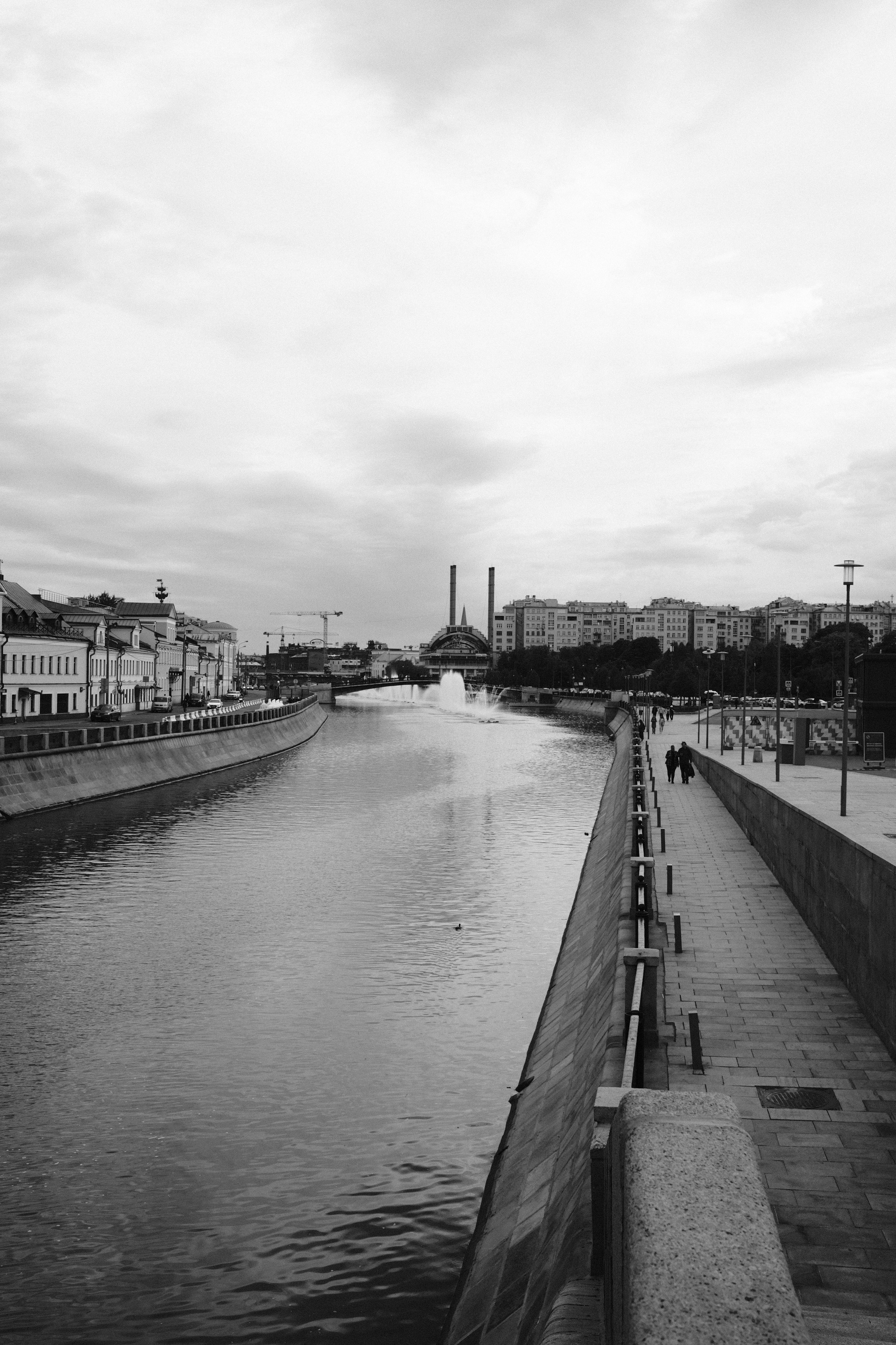 A black and white photo of a city canal with buildings on the left side, a paved walkway on the right, and a bridge in the distance with a fountain. The sky is cloudy, and a few people are walking along the walkway.