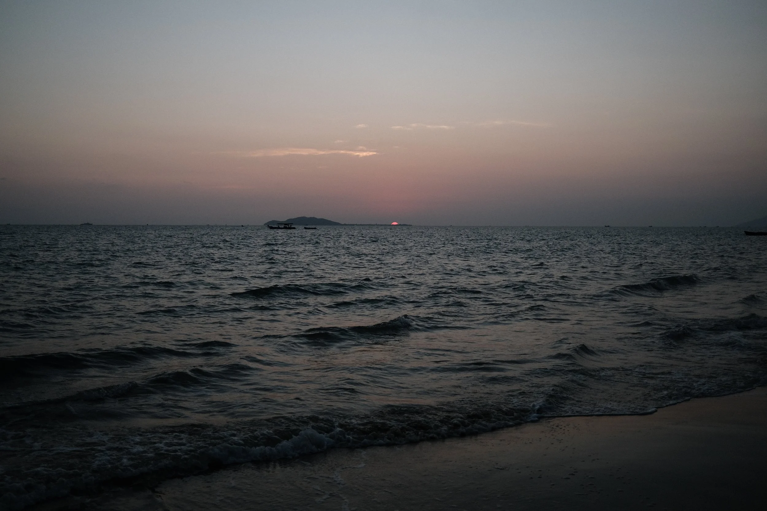 Sunset over the ocean with small waves hitting the shore and boats floating in the distance