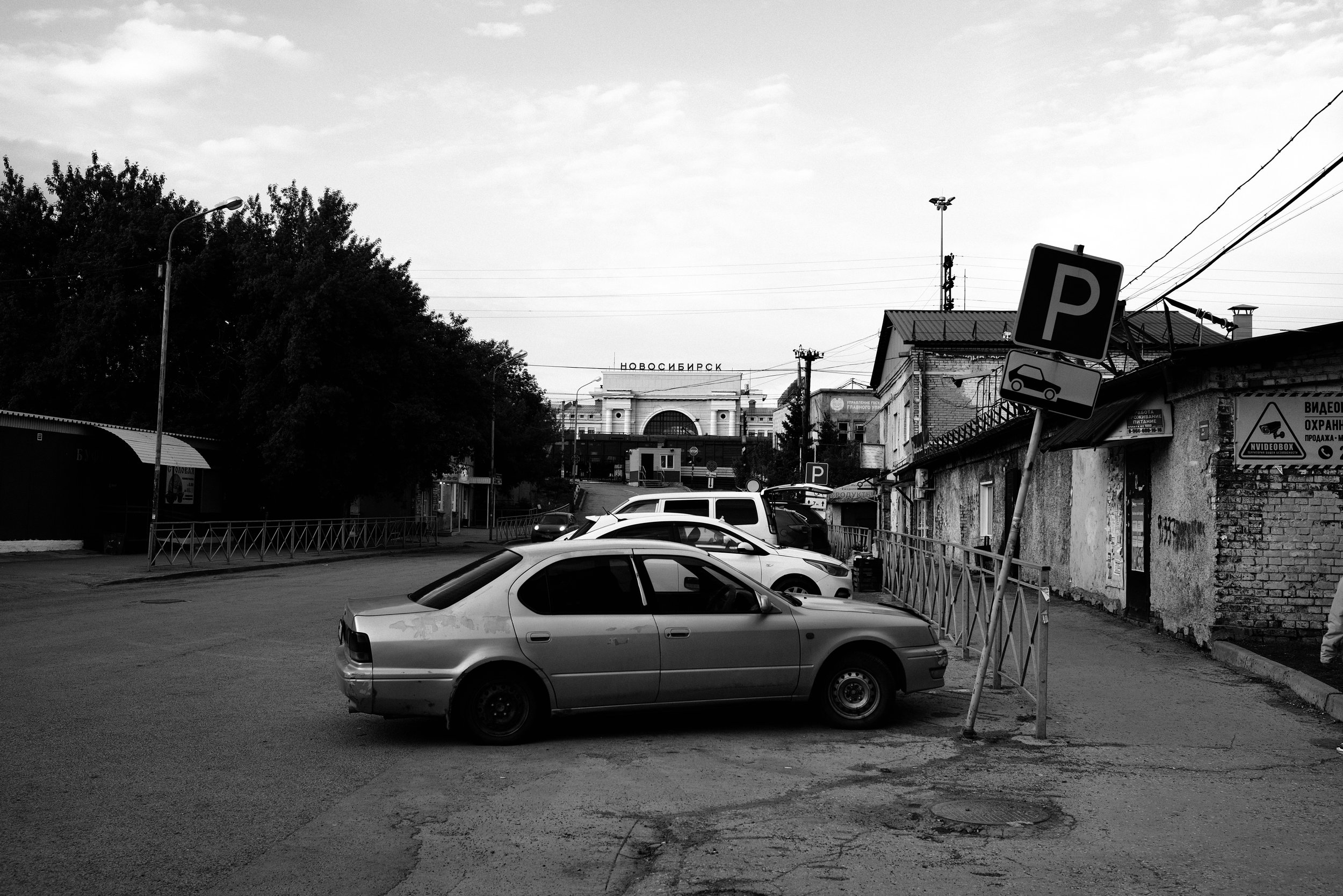 A black and white photo of a parking area with several cars, a parking sign, and a building with the Russian word 'Новосибирск' (Novosibirsk) in the background.
