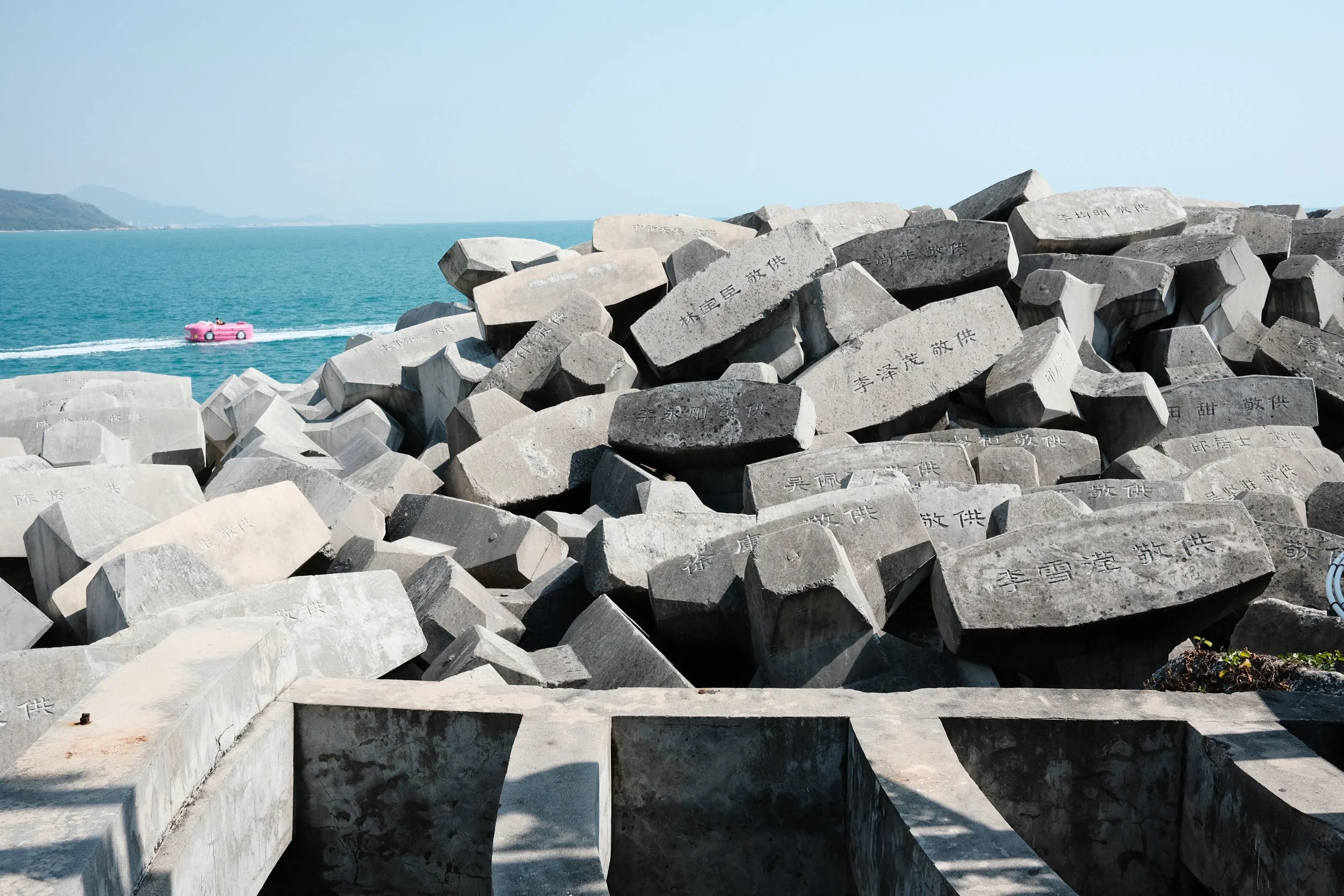 Pile of large gray concrete blocks with Chinese inscriptions, near a body of water with a pink boat in the distance.