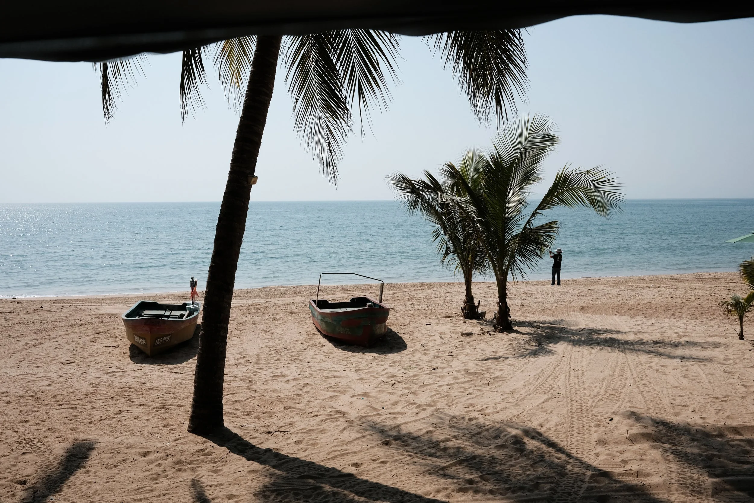 A beach scene with palm trees, two boats on the sand, calm ocean water, and a person taking a photo in the distance.