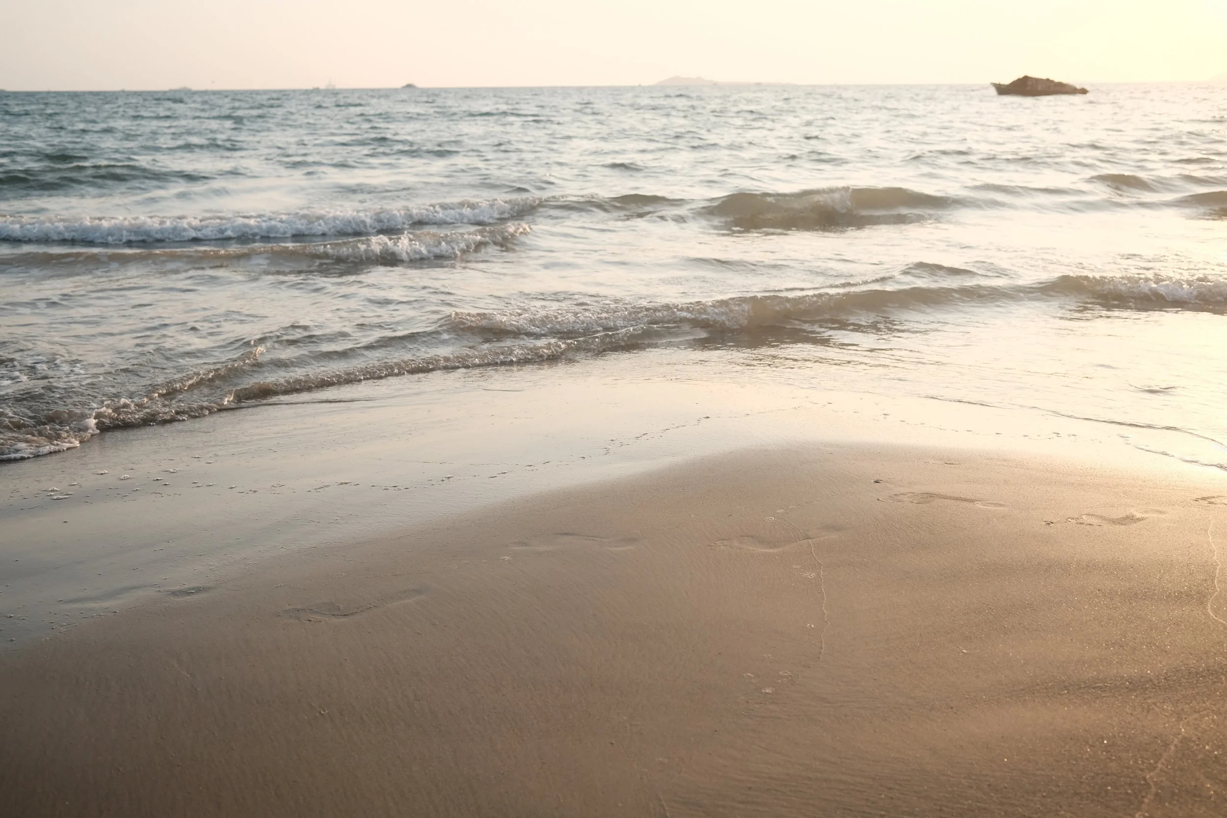 Sunset over a calm ocean, gentle waves rolling onto sandy beach with footprints, distant shoreline, and rocks.