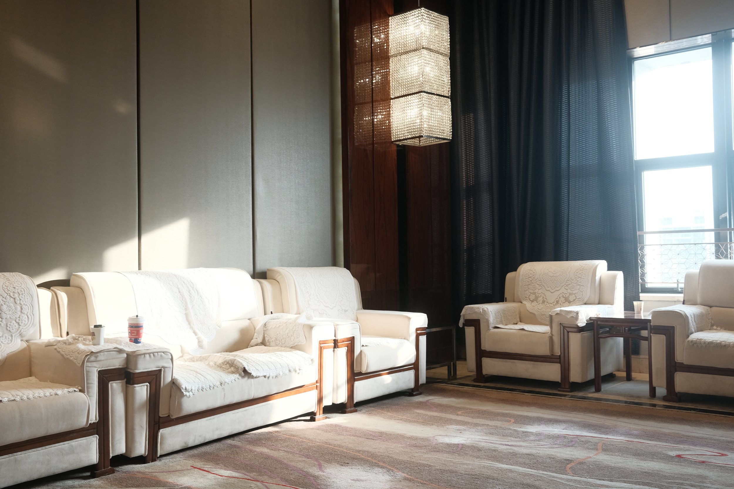 Elegant lounge with white upholstered armchairs and sofas, a large window with black curtains, and a hanging light fixture.