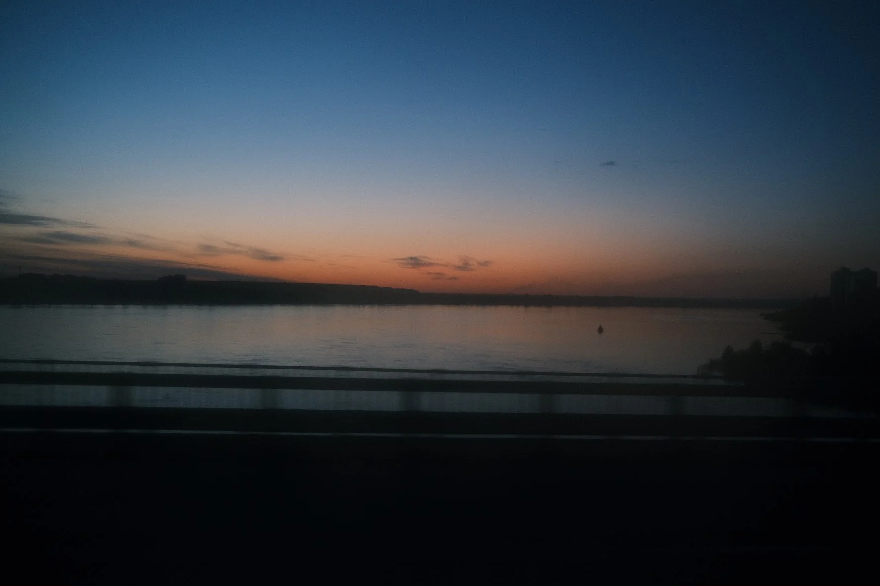 Sunset view over a wide river with a boat, silhouetted trees, and a distant cityscape under a gradient sky transitioning from orange near the horizon to dark blue.