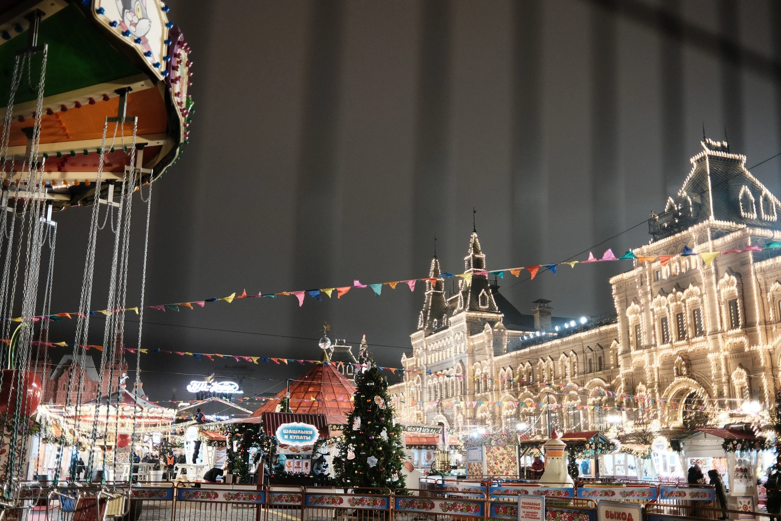 An amusement park at night decorated with colorful lights and flags, featuring a Christmas tree, a carousel, and a grand historic building outlined in bright white lights.