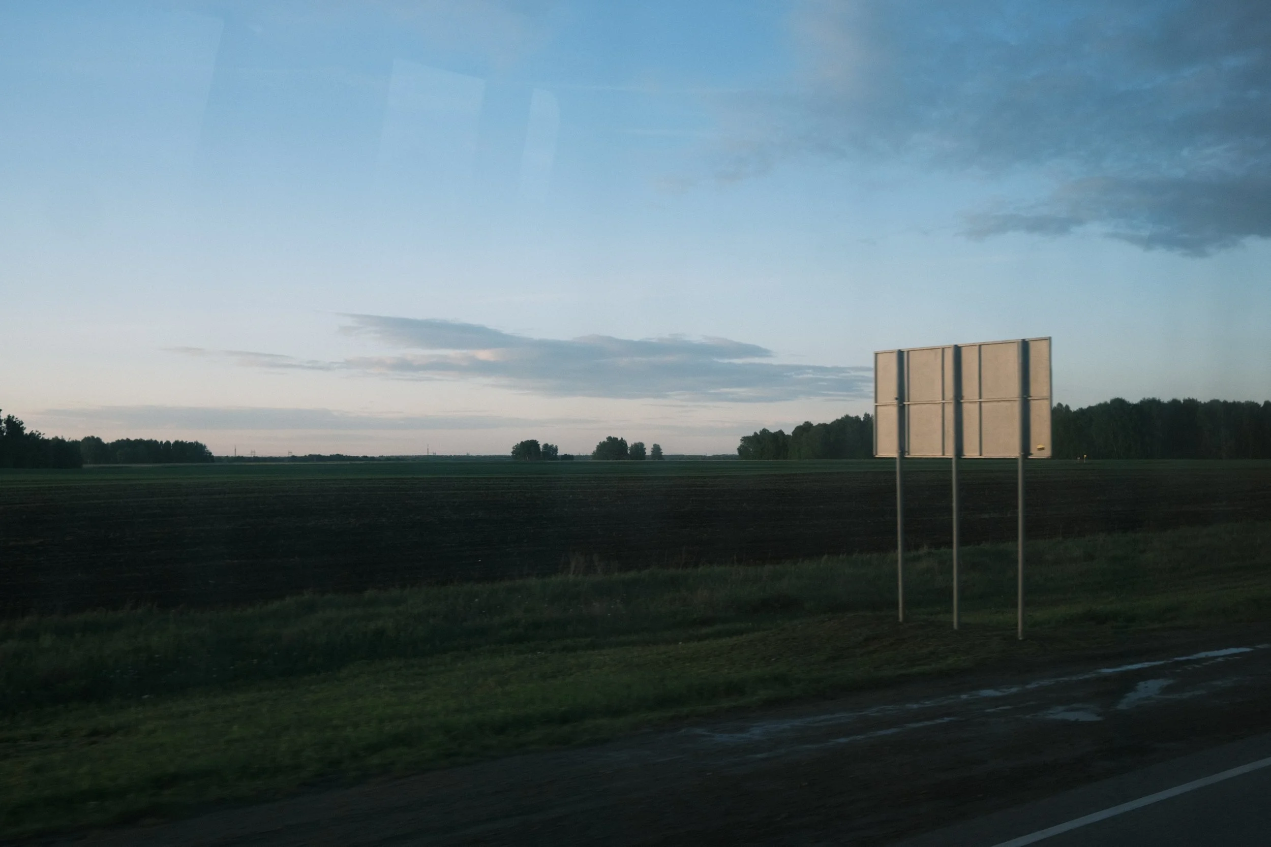 Green fields and a sky with clouds, viewed from a moving vehicle with empty road signs in the foreground.