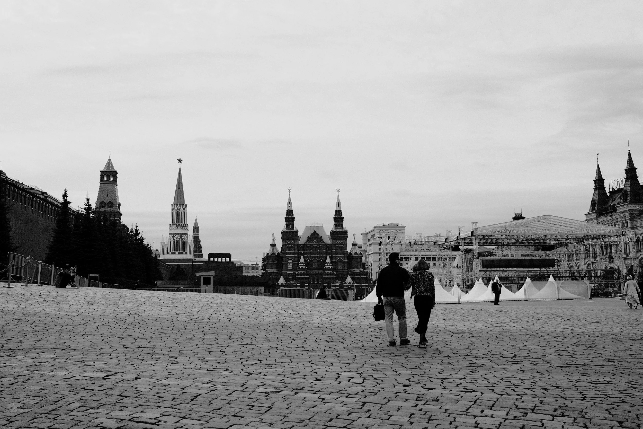 Black and white photo of two people walking hand in hand on a cobblestone street in front of historic Russian architecture, including Kremlin towers and colorful buildings.