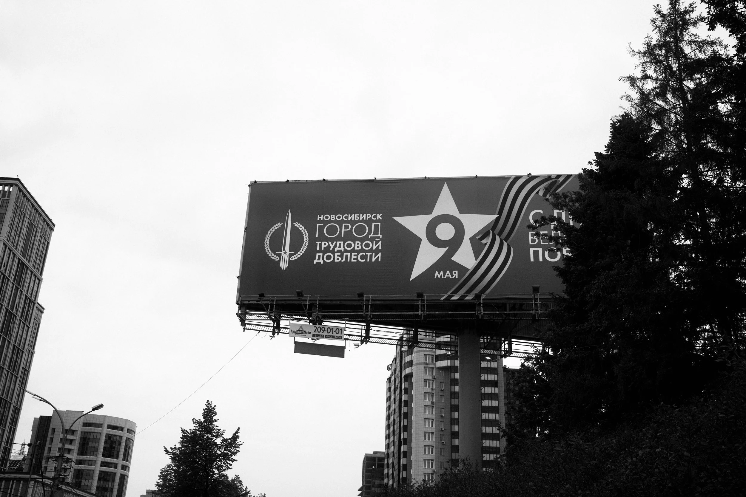 Black and white billboard with Russian text and symbols, celebrating Victory Day on May 9th, in a cityscape with tall buildings and trees.