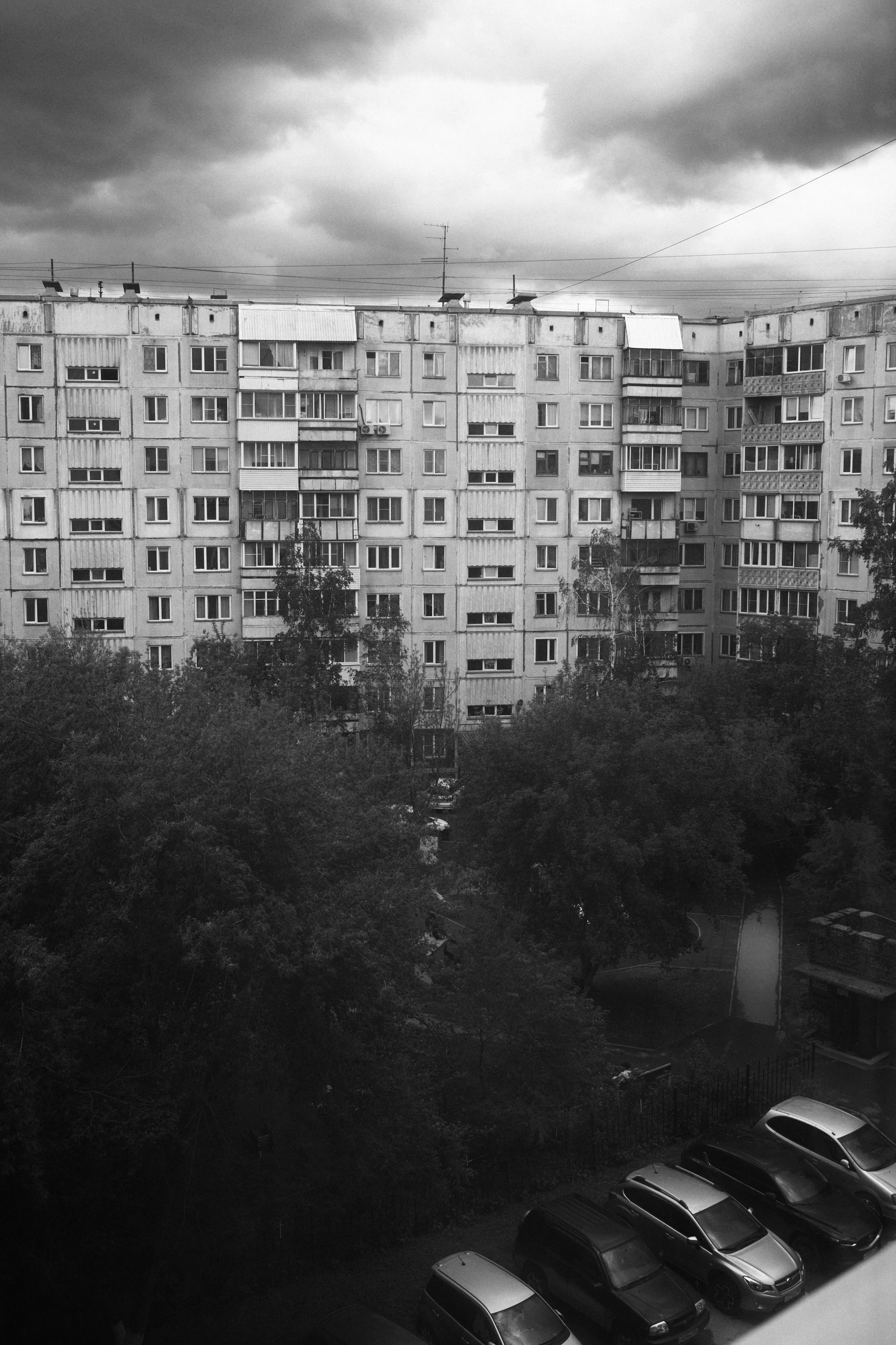 Black and white photo of a high-rise apartment building with multiple stories, surrounded by trees and parked cars at the bottom.