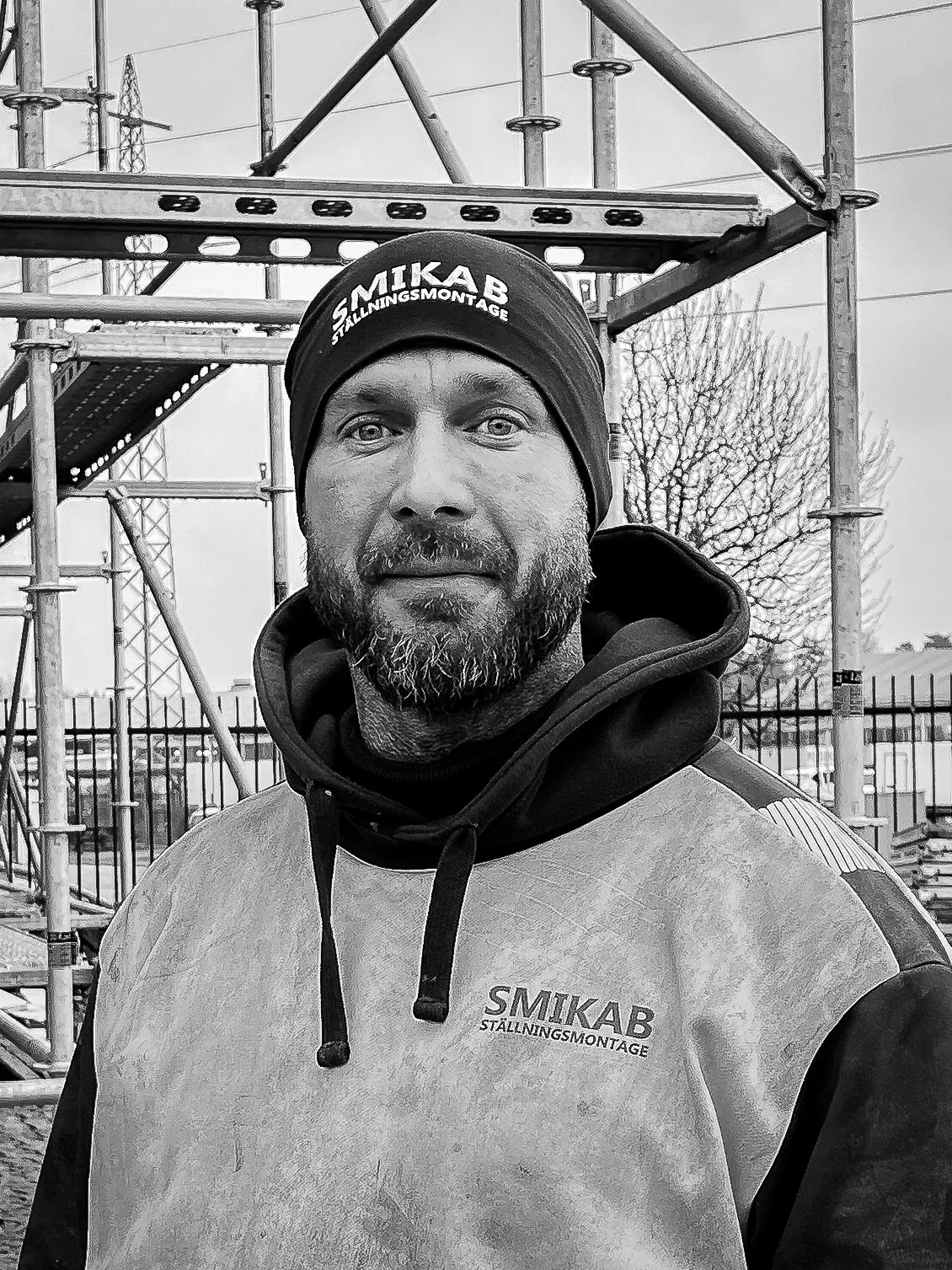 En man med skägg och keps, klädd i arbetskläder med texten 'SMIKAB STÄLLNINGSMONTAGE', står framför en byggnadsställning.