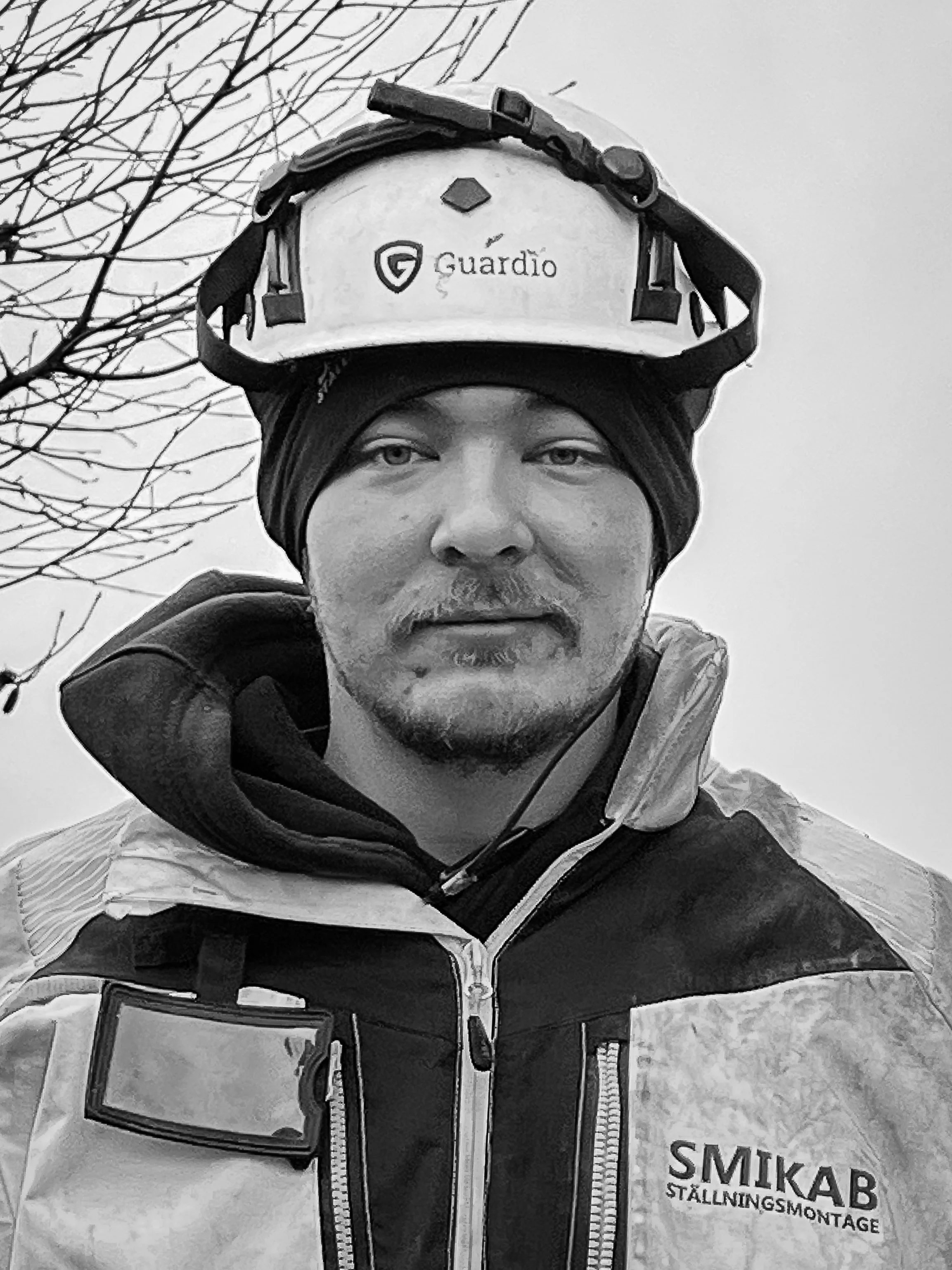 Man i arbetskläder med hjälm och skyddsutrustning, stående utomhus, med träd i bakgrunden.