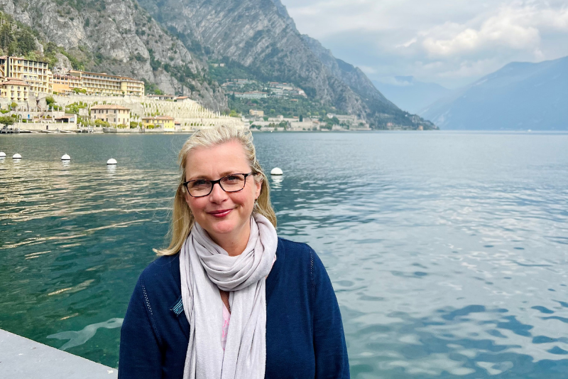 Sarah tanding by Lake Garda in Italy with mountains and lakeside buildings in the background, calm water and cloudy sky, capturing a peaceful solo travel moment in northern Italy.