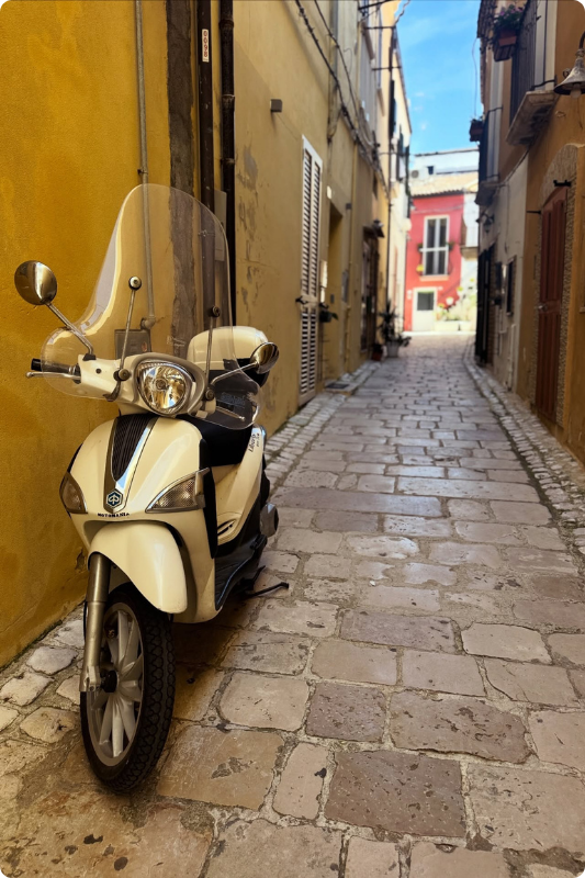Classic white Italian scooter parked along a narrow cobbled street in a charming historic village, framed by warm ochre walls and colourful buildings, capturing the timeless beauty of authentic slow travel in Italy.