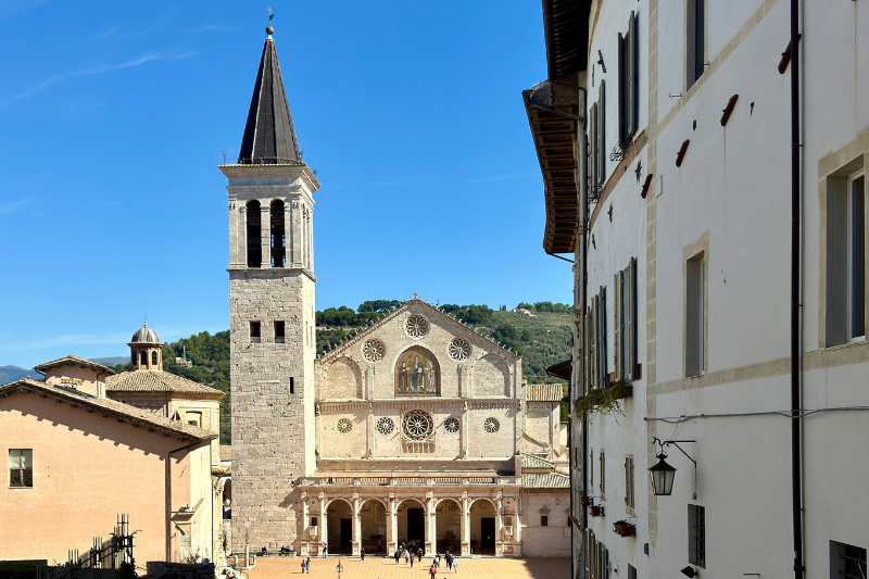 Spoleto Cathedral (Duomo di Spoleto) in Umbria, Italy, with bell tower and Romanesque façade in a sunlit piazza surrounded by historic buildings and blue sky.