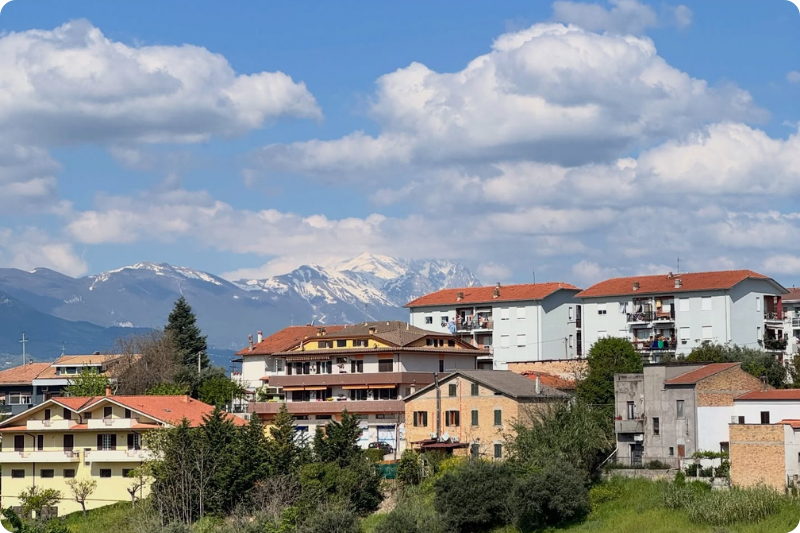 Scenic Italian hillside village with terracotta rooftops, rolling green countryside, olive groves and snow-capped mountains under a bright blue sky, showcasing authentic rural Italy and slow travel landscapes.