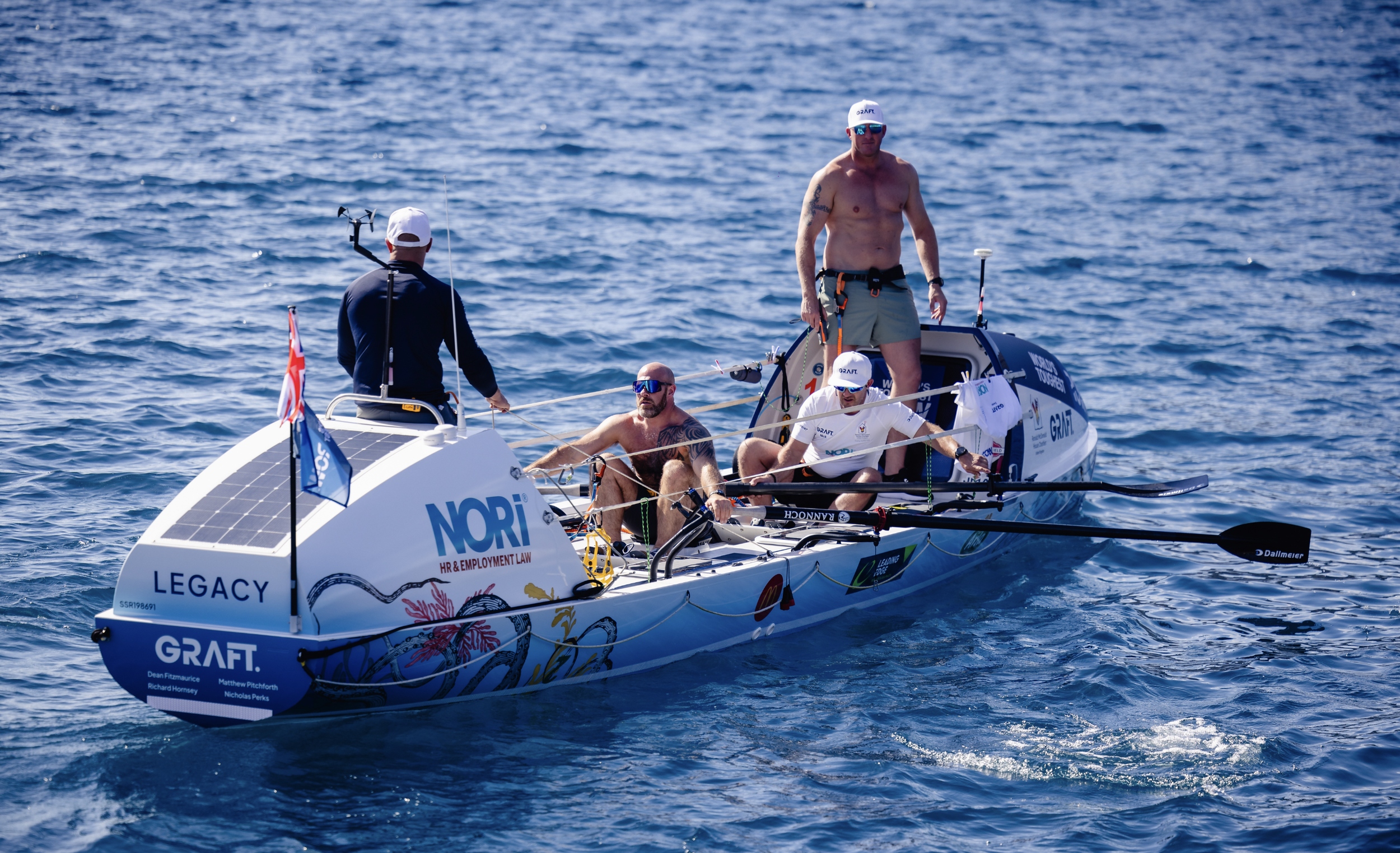 A group of four men on a small racing sailboat on the water, with one standing shirtless and others seated, all wearing caps and sunglasses, with various sponsorship logos on the boat.
