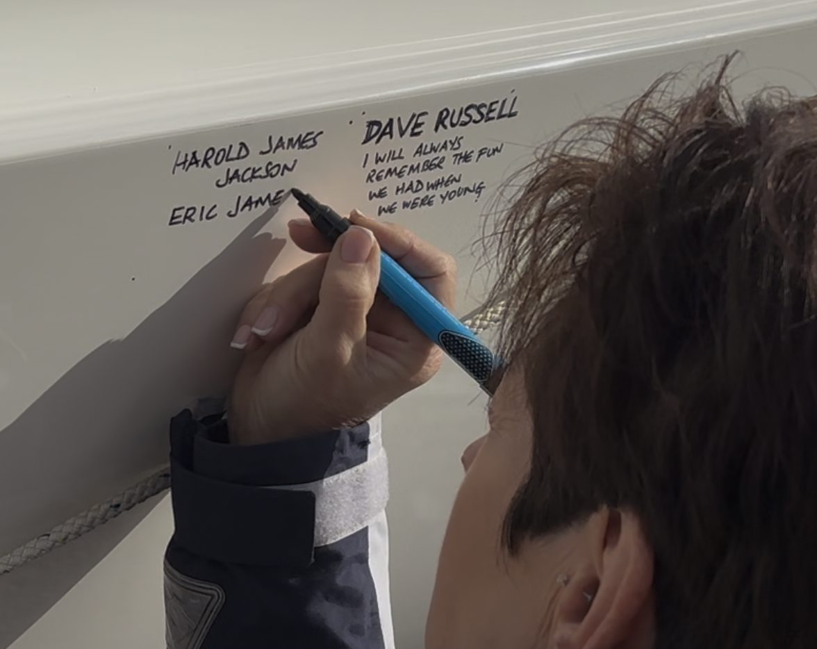 Person writes names and a message on a whiteboard with a blue marker, with a focus on the person's hand and the writing.