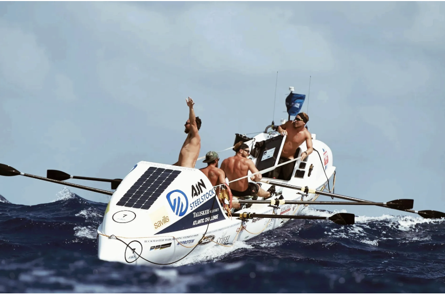 Four men rowing a white sailboat in the ocean with solar panels on its deck, under a cloudy sky.