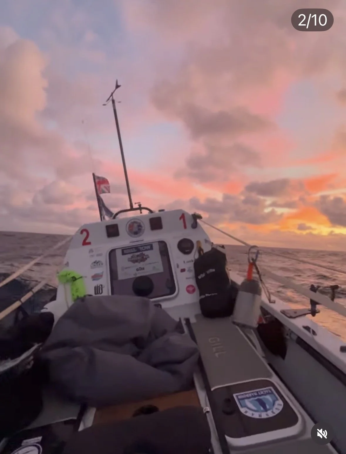View from a boat at sunset or sunrise with pink clouds and calm water, showing fishing equipment, a GPS device, and a weather-resistant bag on the boat.