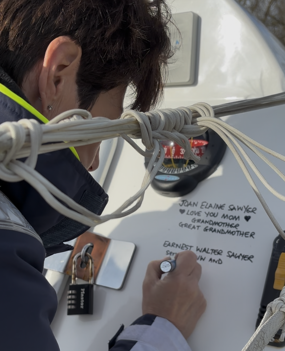 A person writing on a whiteboard or sign, with handwritten messages of love and remembrance for family members, including Joan Elaine Sawyer and Ernest Walter Salter, surrounded by ropes on a boat.