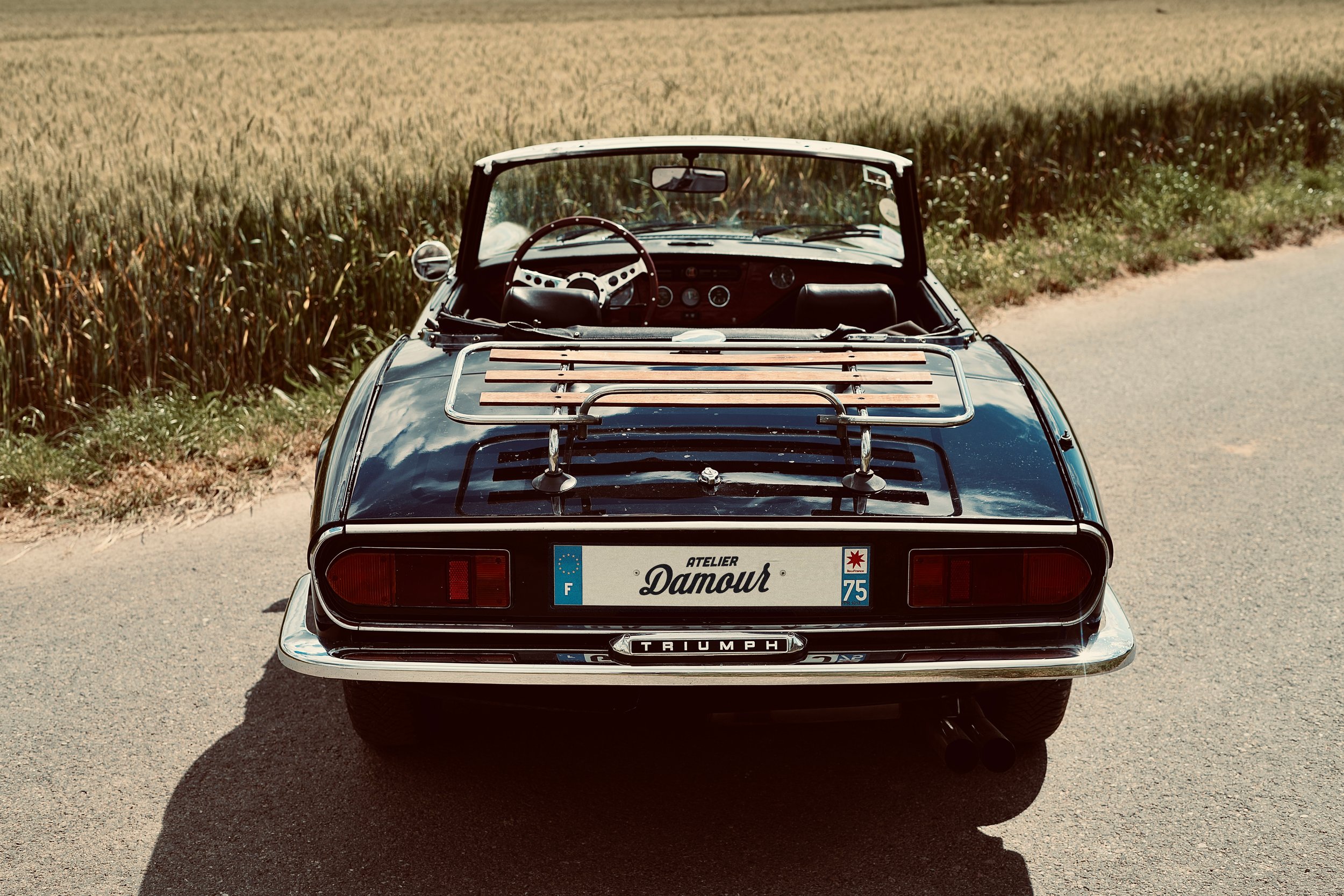 Voiture décapotable rétro Triumph, vue de dos, sur une route de campagne, près d'un champ. Plaque d'immatriculation indique "Atelier Damour". Un porte-bagages est fixé à l'arrière.