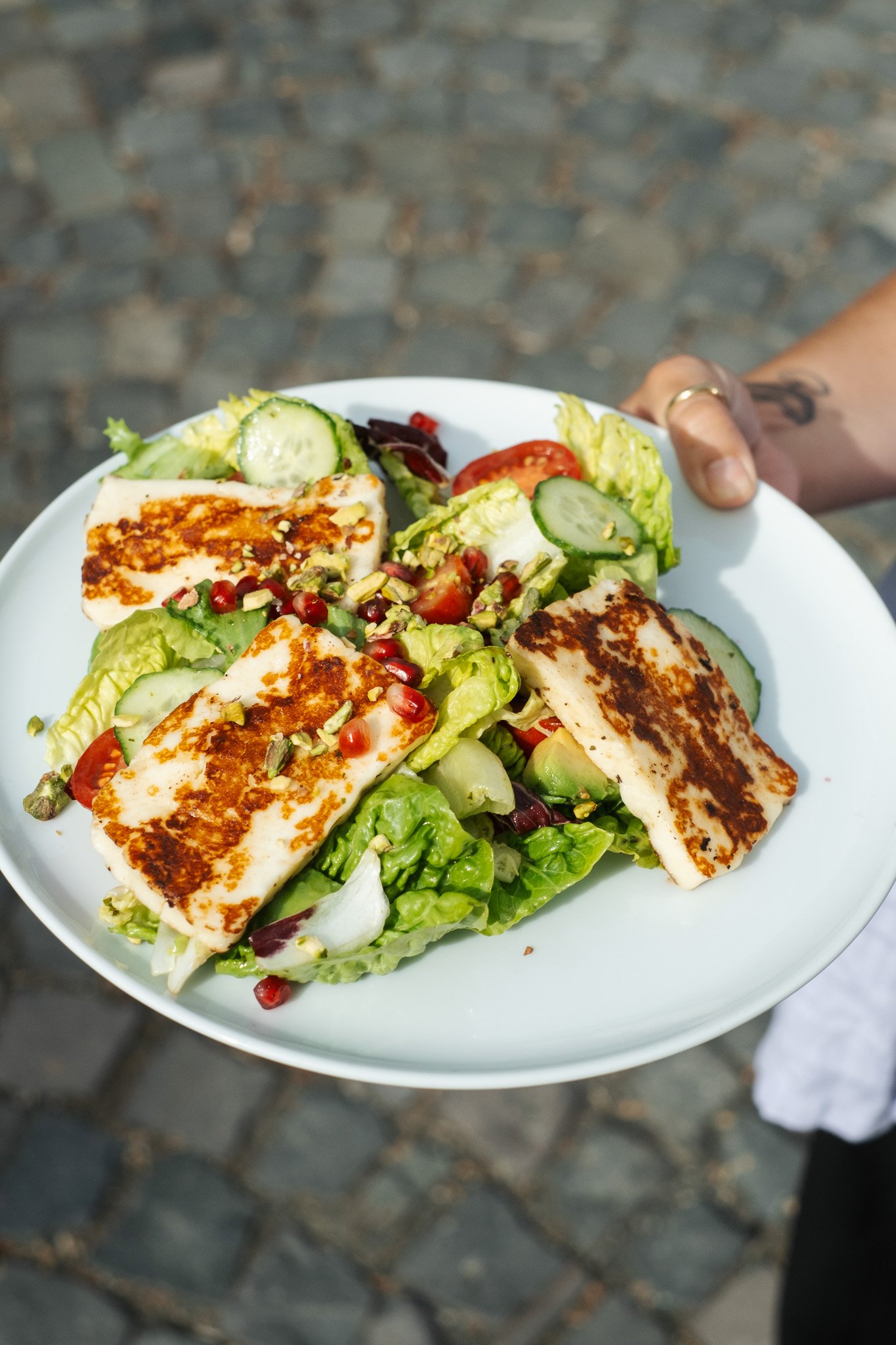 A white plate of grilled halloumi cheese served over a bed of fresh salad greens, cherry tomatoes, cucumber slices, chopped nuts, and pomegranate seeds, held by a person with a tattoo on their arm, standing on a cobblestone surface.
