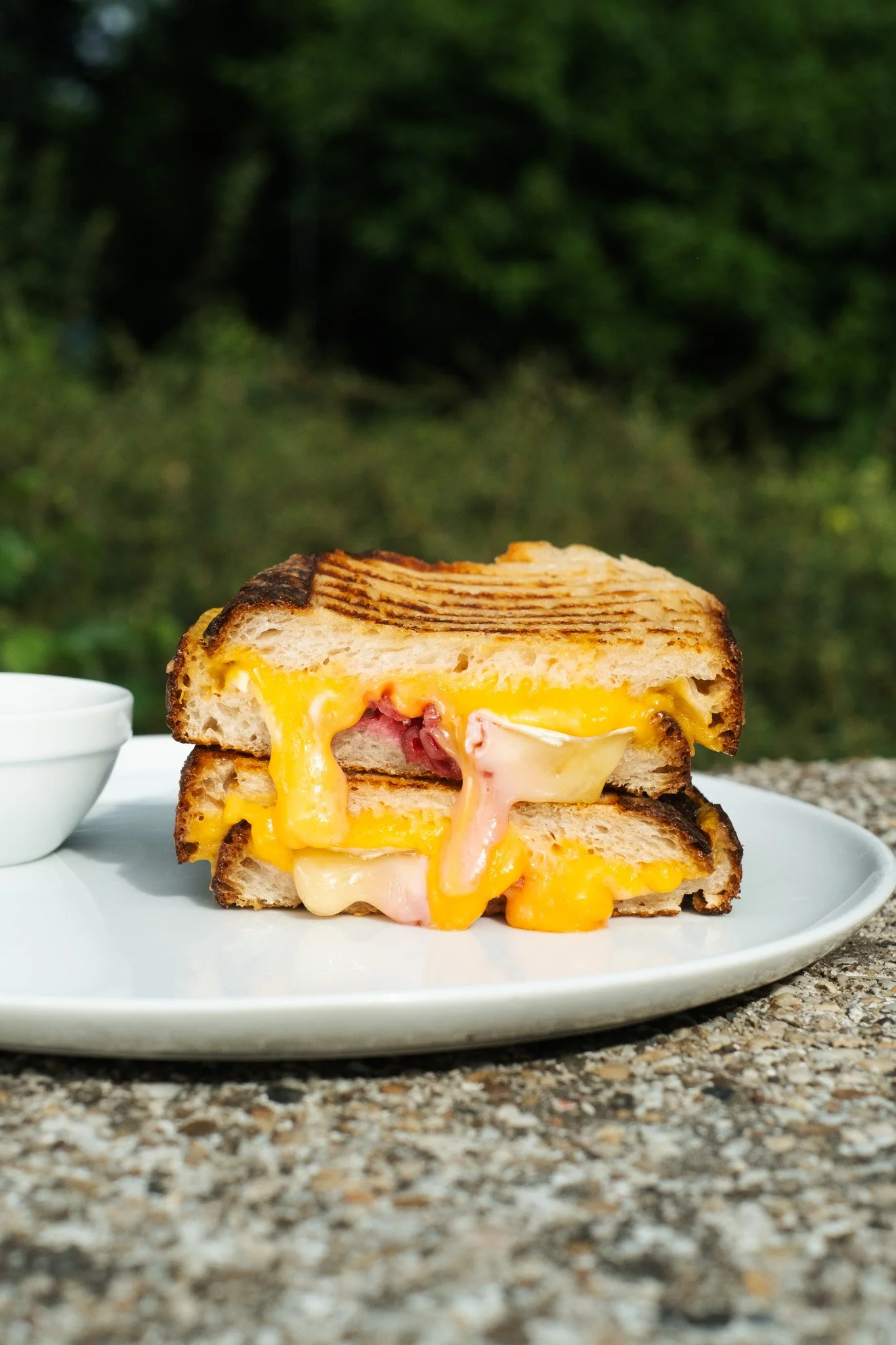 Close-up of a grilled breakfast sandwich with melted cheese, sliced ham, and grilled bread on a white plate outdoors, with a blurred green background.