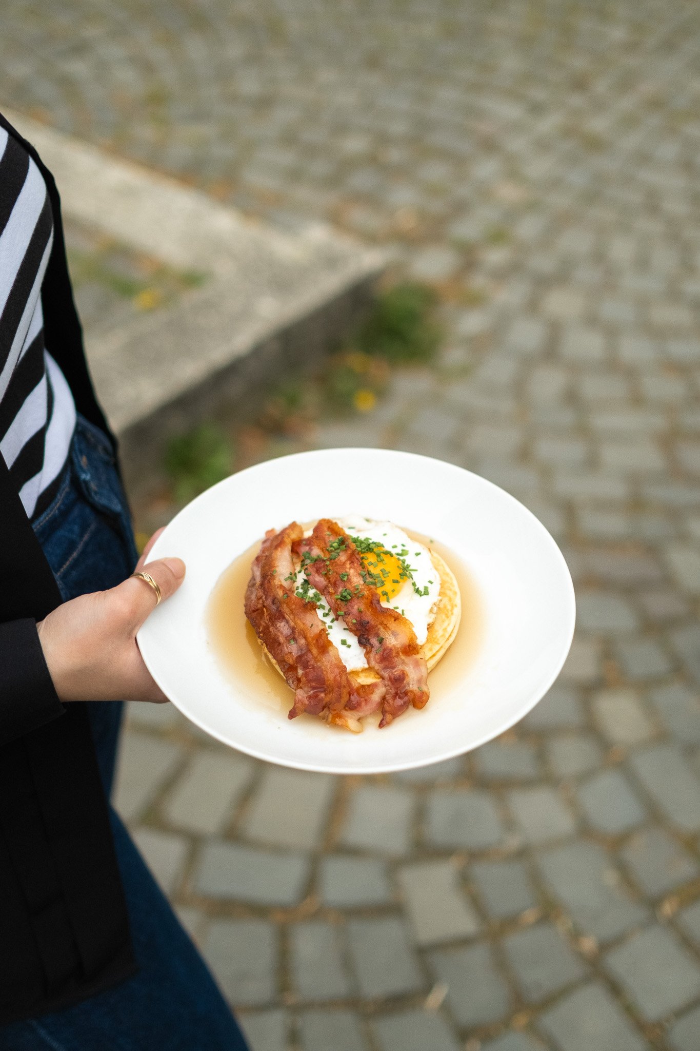 A person holding a white plate with a breakfast dish that includes an egg, bacon, and some garnish. The person is wearing a black and white striped shirt and a ring on the finger of their left hand. The background shows cobblestone pavement.
