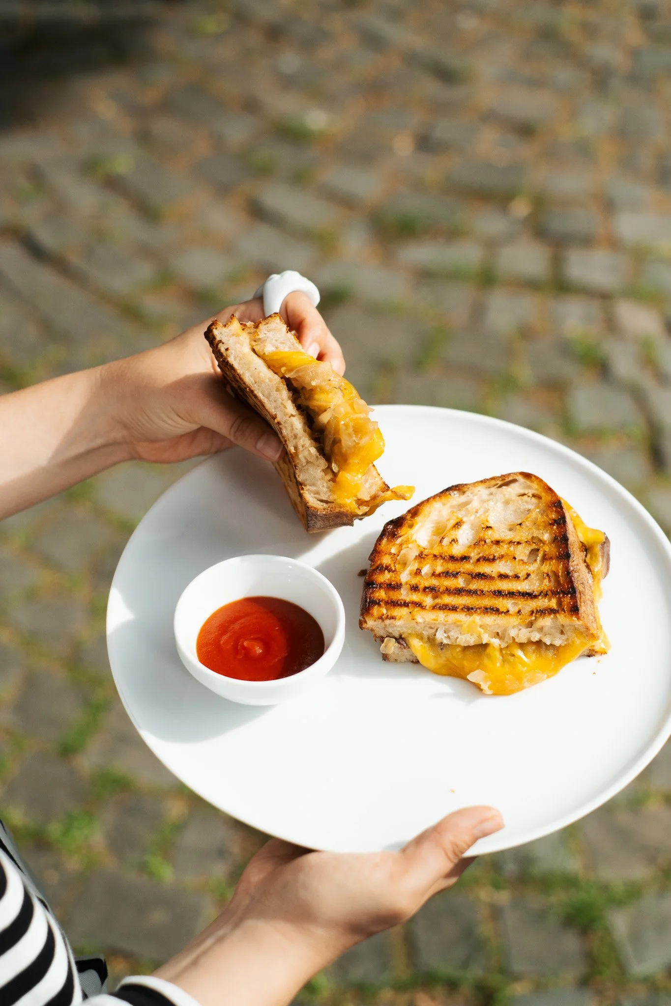 Person holding a white plate with a grilled sandwich and a small bowl of ketchup, outdoors on a fall day.