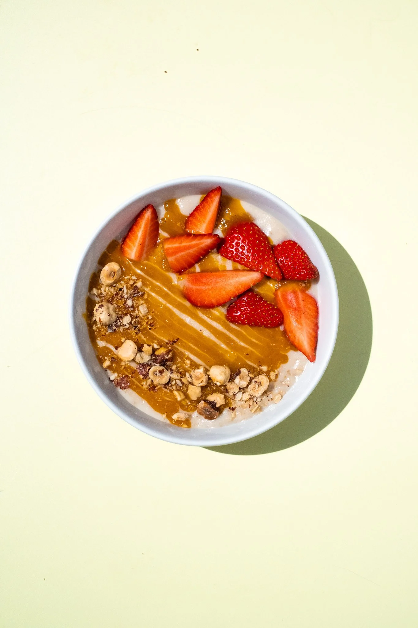 A bowl of yogurt topped with sliced strawberries, chopped nuts, caramel sauce, and granola on the side, placed on a light green surface.
