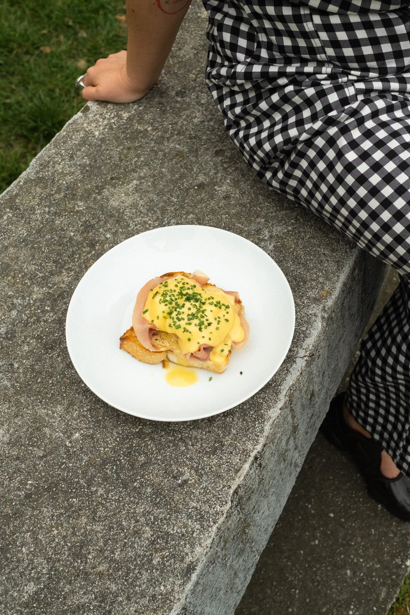 A plate of eggs Benedict with ham and chopped chives, topped with hollandaise sauce, placed on a textured concrete surface outdoors.