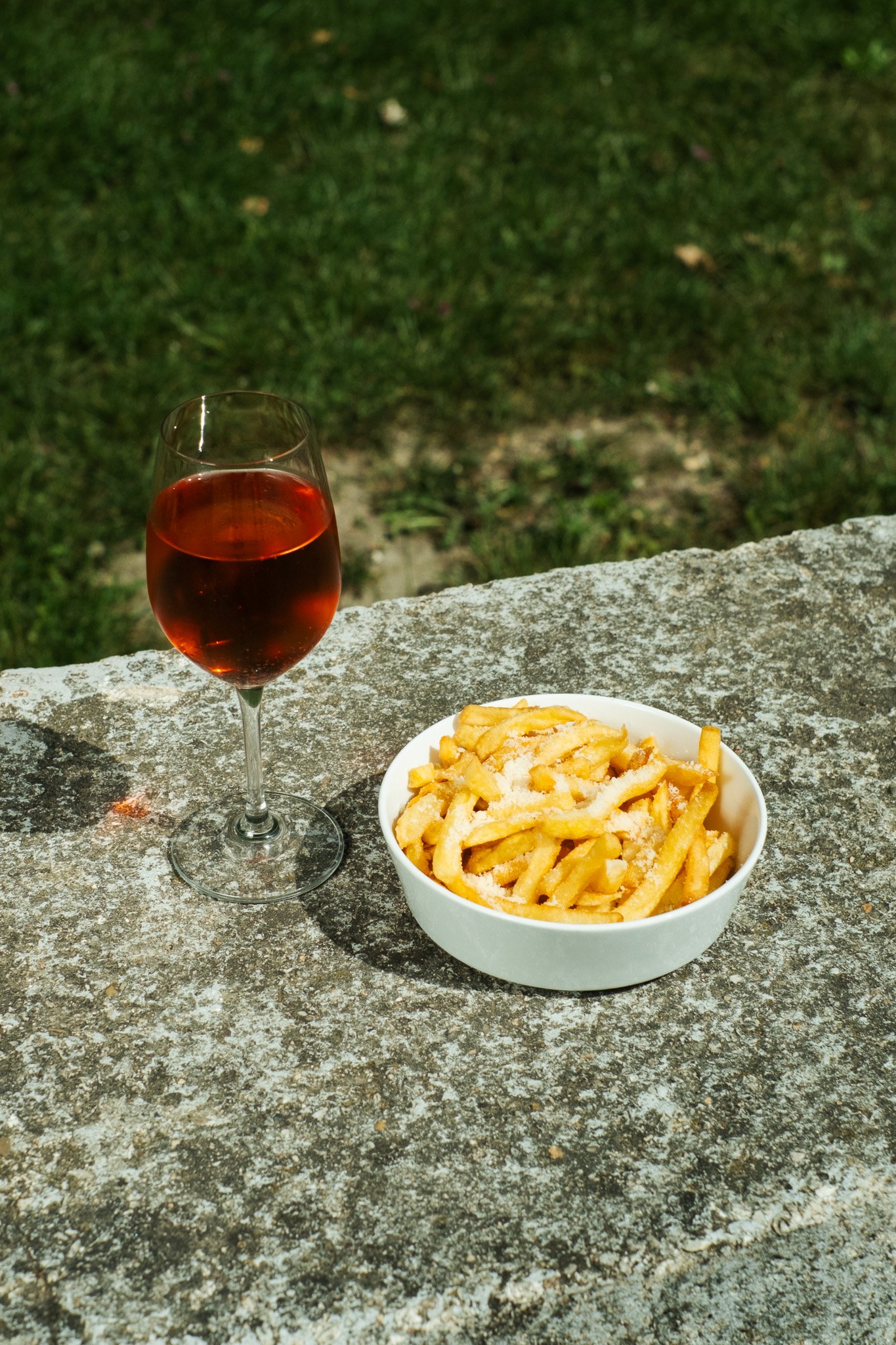 A glass of red wine and a bowl of cooked pasta with grated cheese on top, placed on a stone surface outdoors with green grass in the background.
