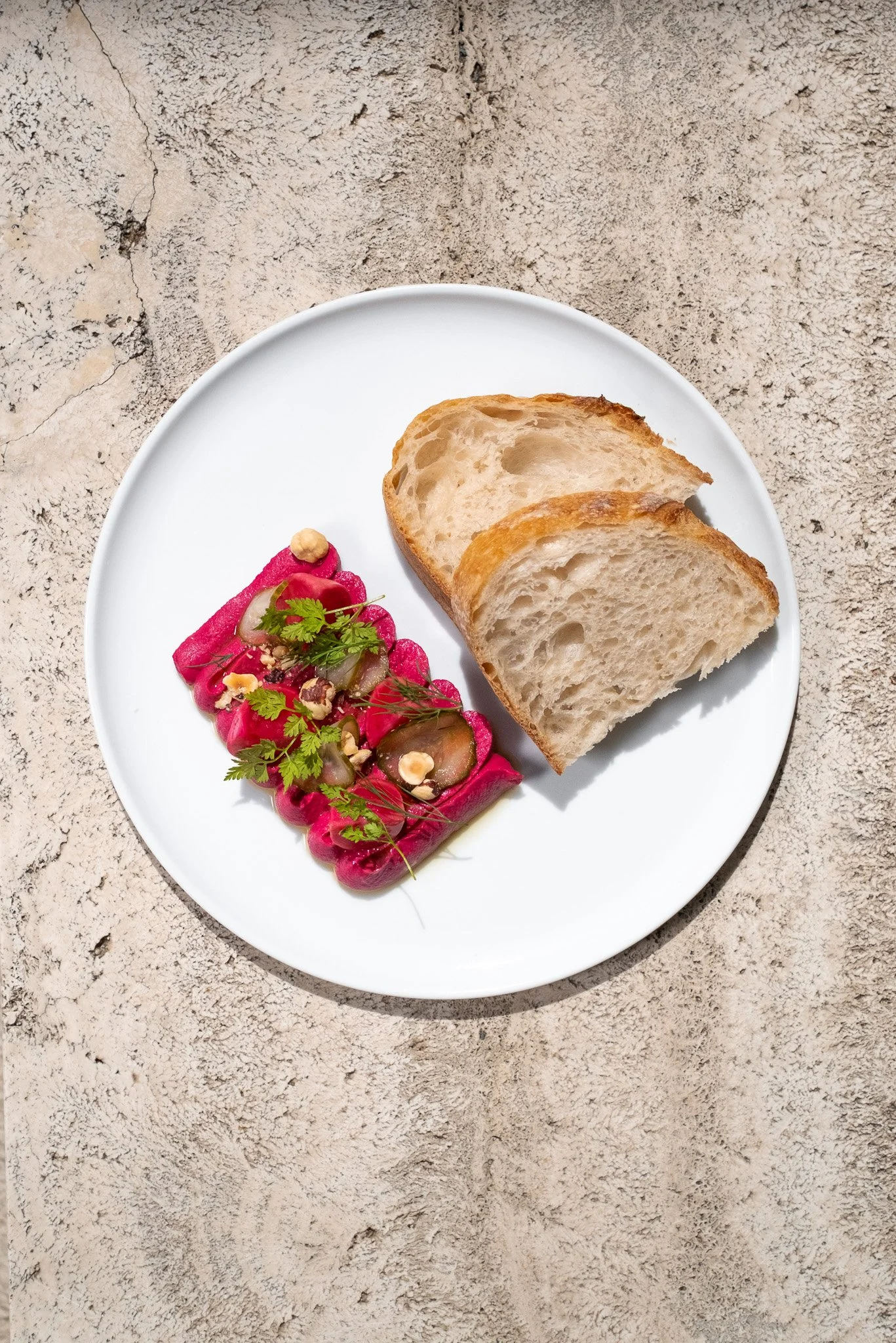 A white plate with two slices of bread and a small salad with pink pickled vegetables, garnished with herbs and nuts, on a textured surface.