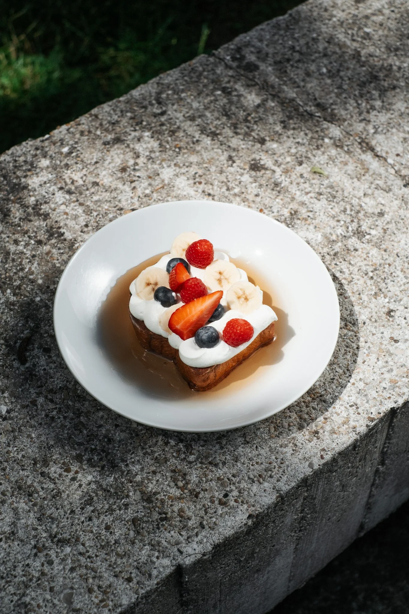 A slice of cake topped with whipped cream and fresh berries, including strawberries, blueberries, bananas, and raspberries, on a white plate on a stone surface outdoors.