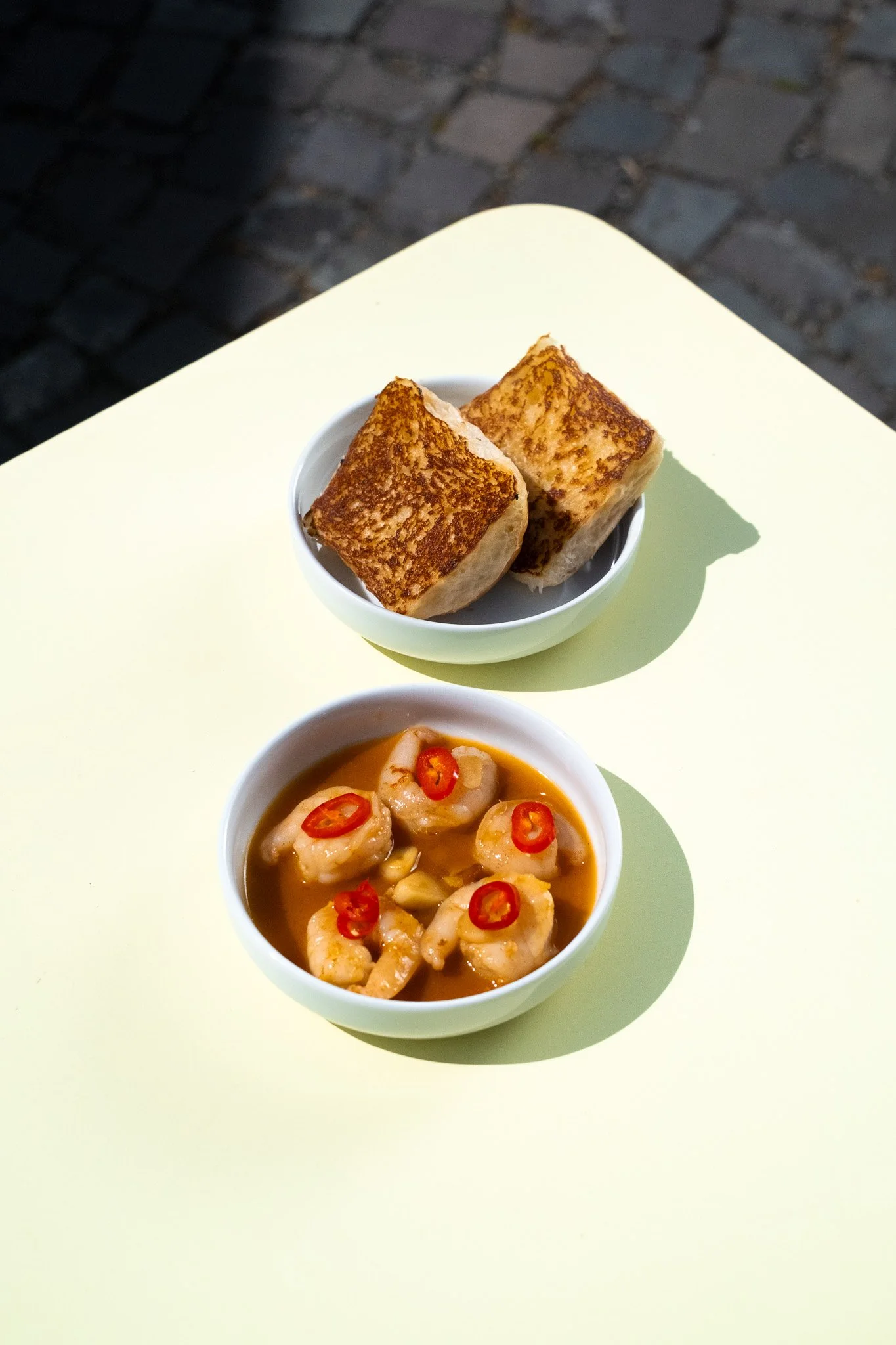 Two bread rolls in a small white bowl placed on a light-colored table, with a dish of shrimp in sauce garnished with red chili slices in front of it, outdoors on a cobblestone surface.
