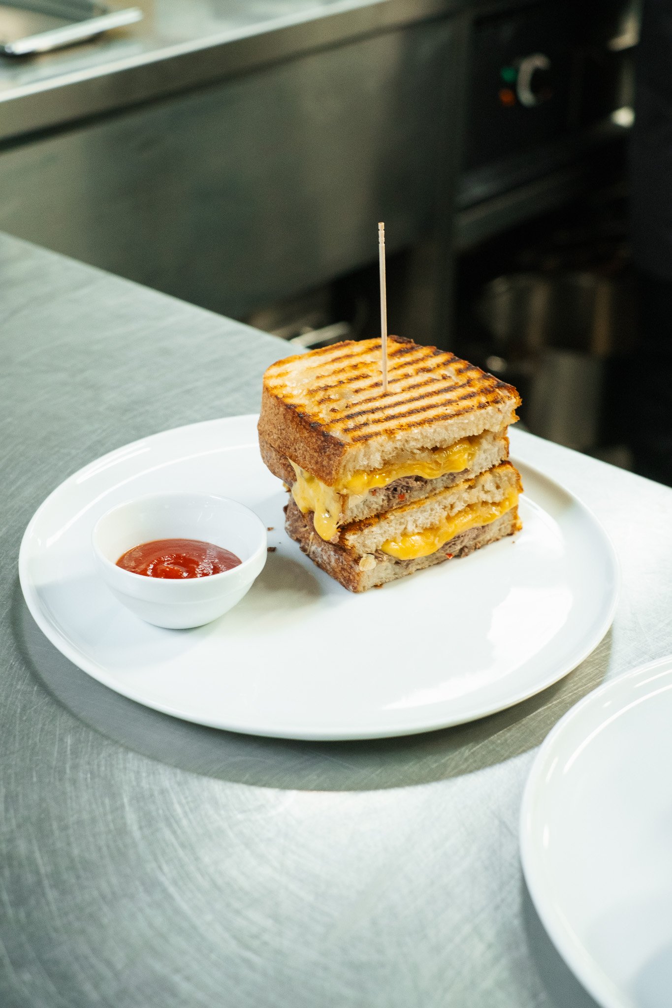 A grilled cheese and hamburger patty sandwich on a white plate with a small cup of ketchup.