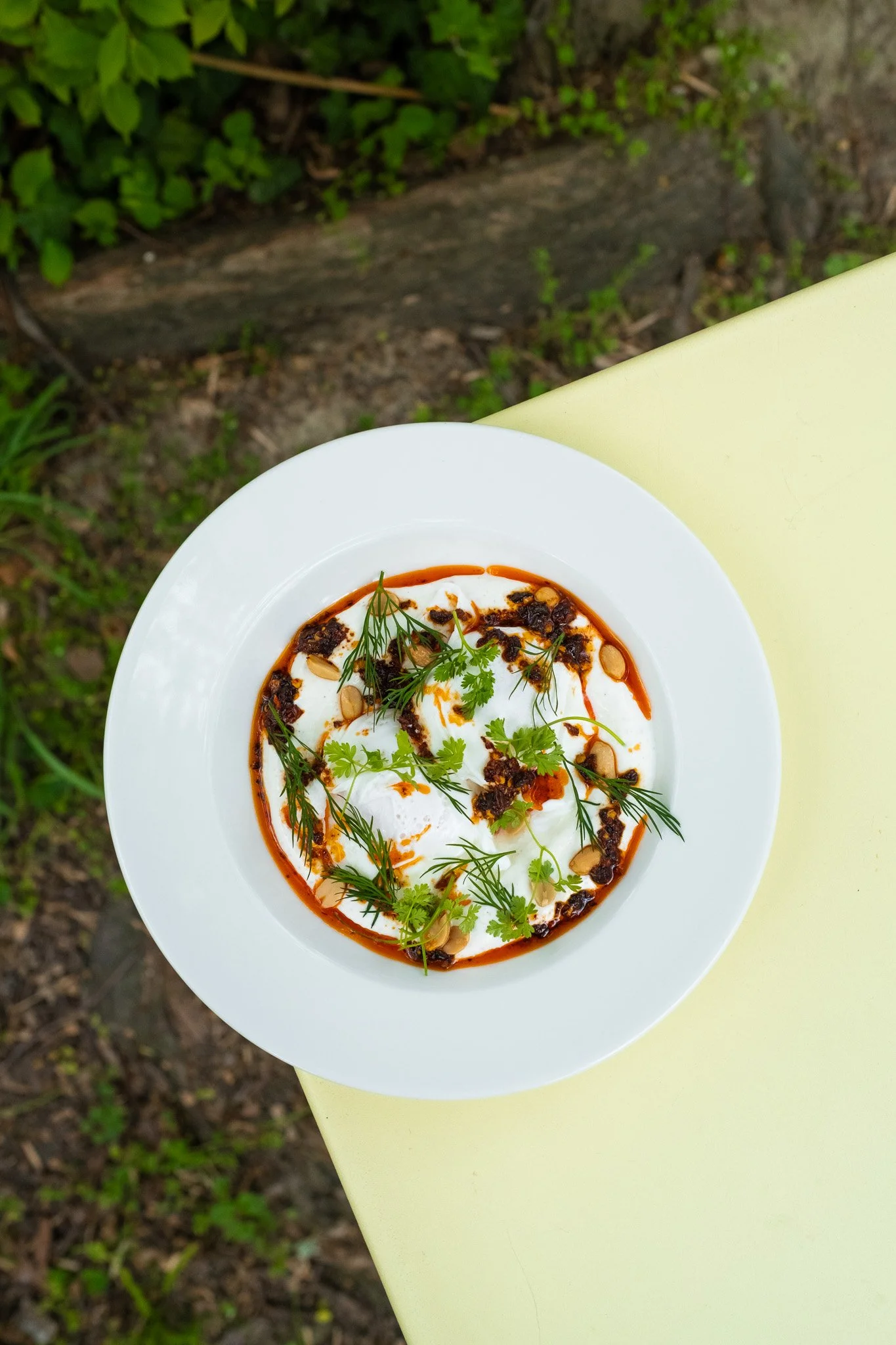 A bowl of yogurt garnished with herbs, nuts, and a spicy sauce on a yellow table outdoors.