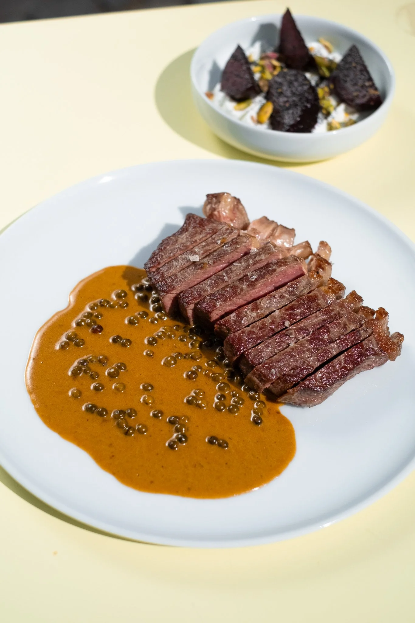 A plate with sliced steak topped with a brown sauce and peppercorns. In the background, a small bowl of whipped cream with dark purple chips and chopped nuts.