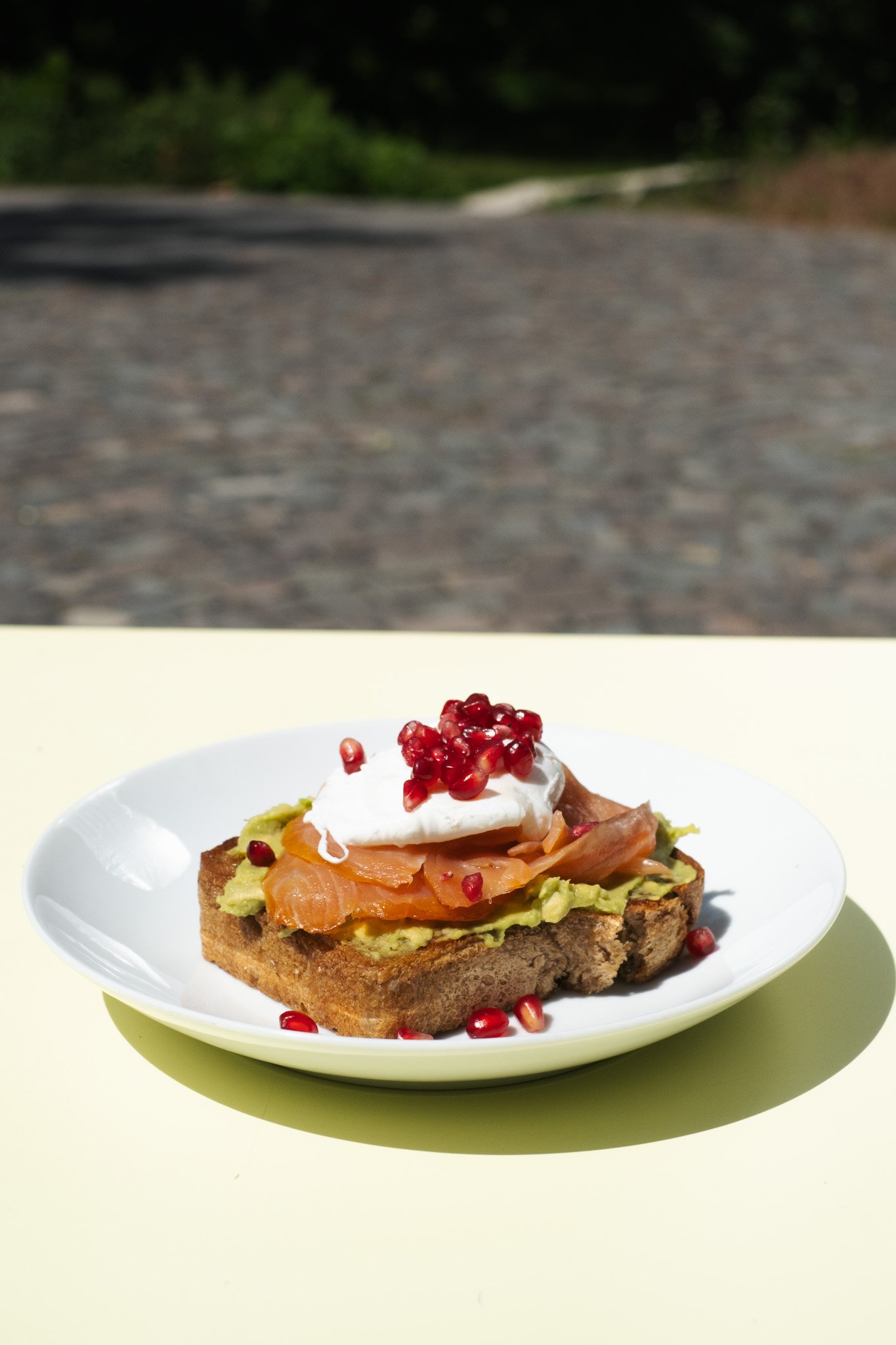 Open-faced flatbread topped with avocado, smoked salmon, a dollop of sour cream or yogurt, pomegranate seeds, and chopped herbs, placed on a white plate.