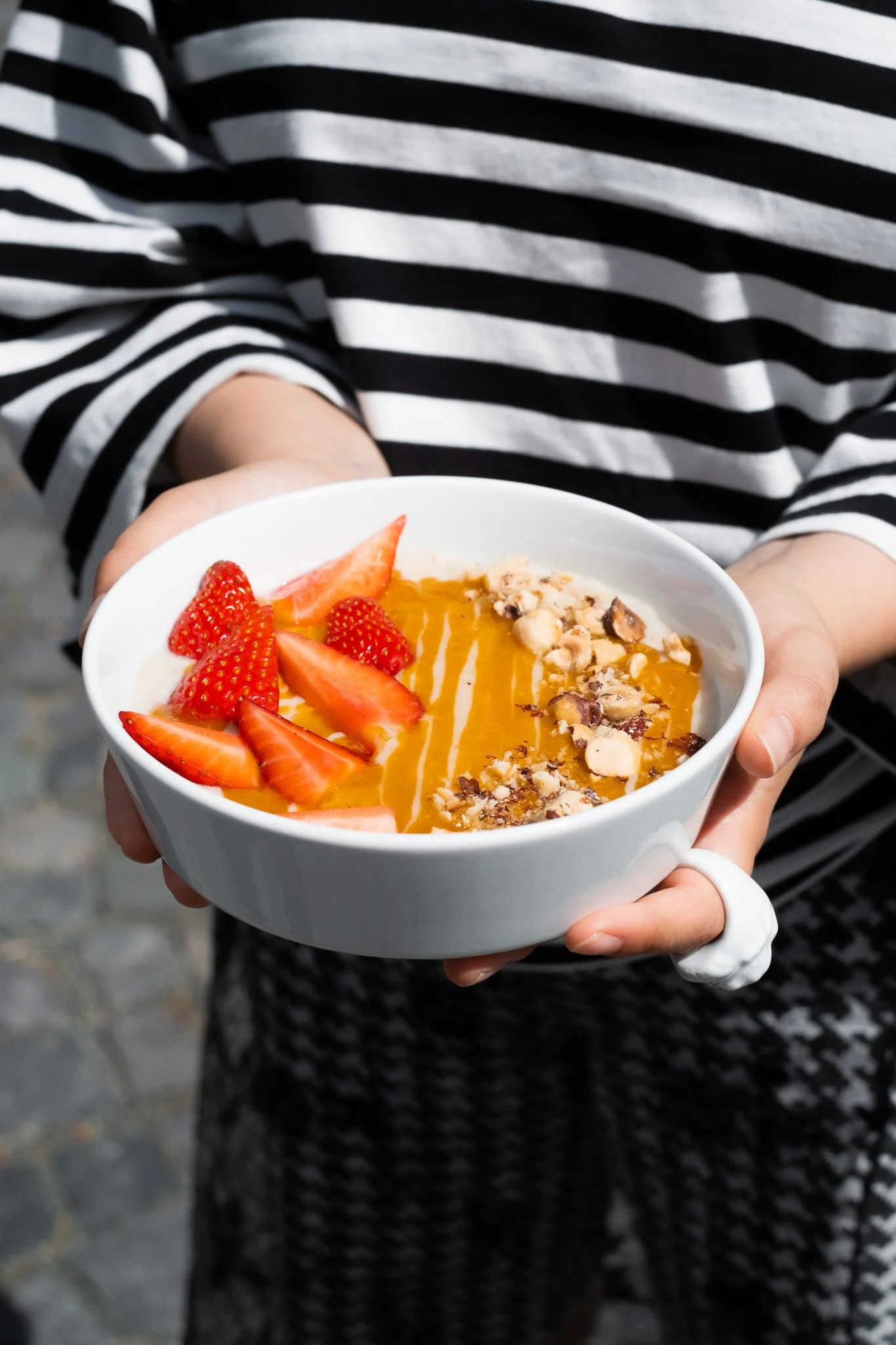 Person wearing a black and white striped shirt holding a bowl of smoothie topped with strawberries, chopped nuts, and honey.