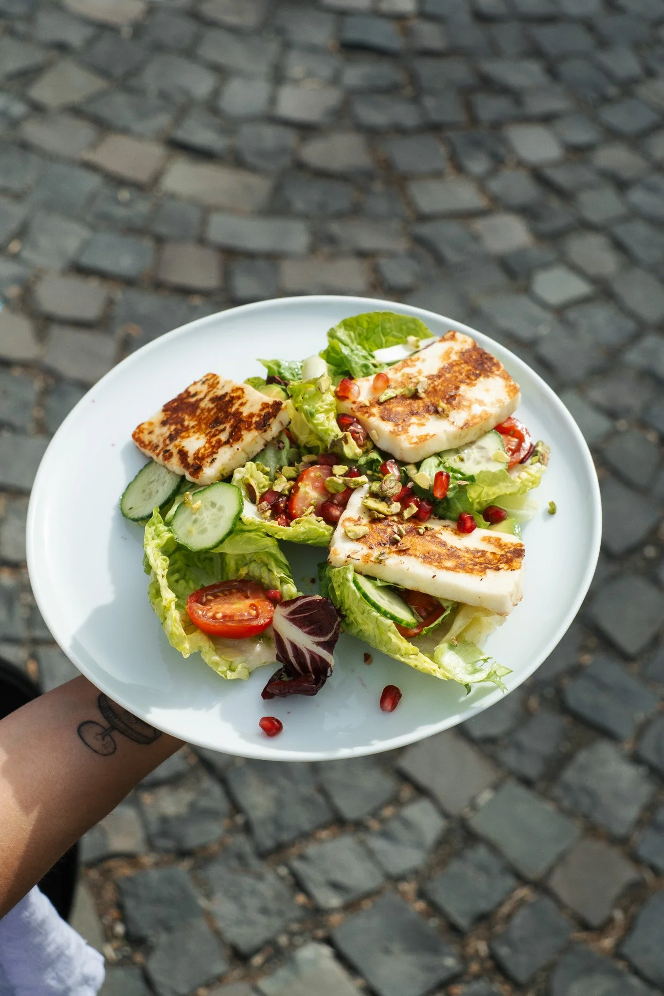 A white plate with grilled halloumi cheese, cucumber slices, cherry tomatoes, pomegranate seeds, and mixed salad greens being held outdoors over a cobblestone street.