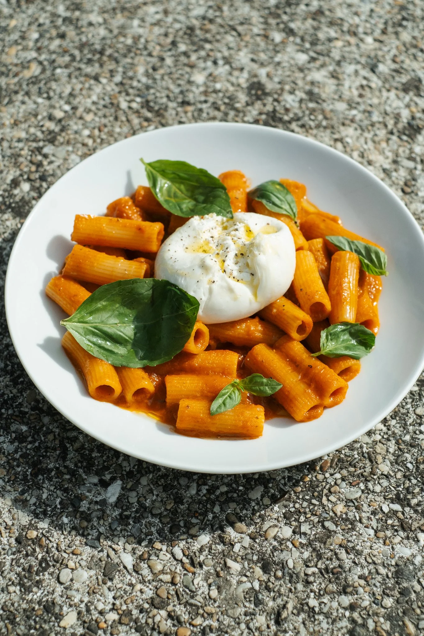 A white bowl contains rigatoni pasta in tomato sauce, garnished with basil leaves, topped with a poached egg, on a gravel surface.