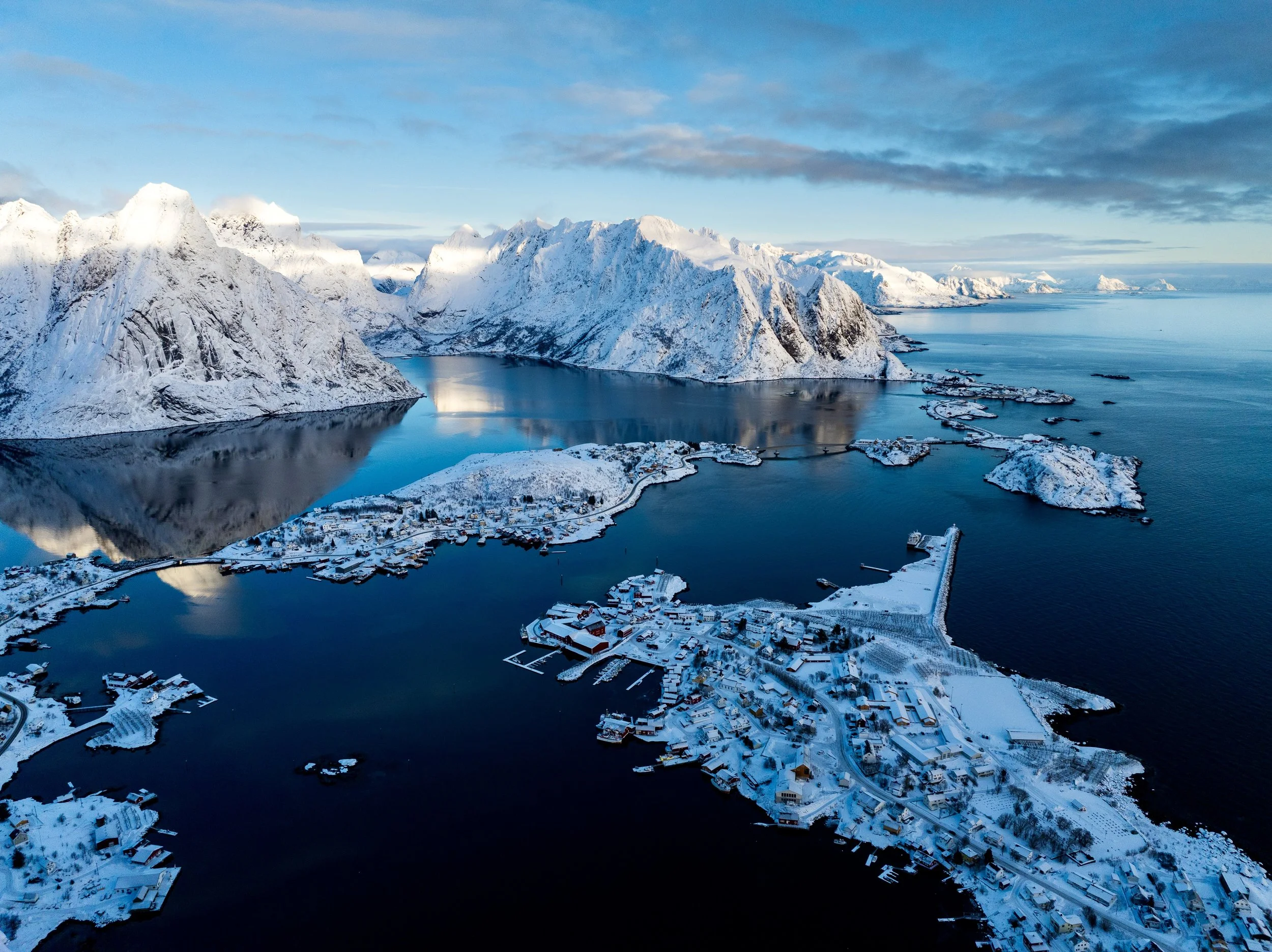A snowy fjord landscape with snow-covered mountains, a small village, and boats in the water.