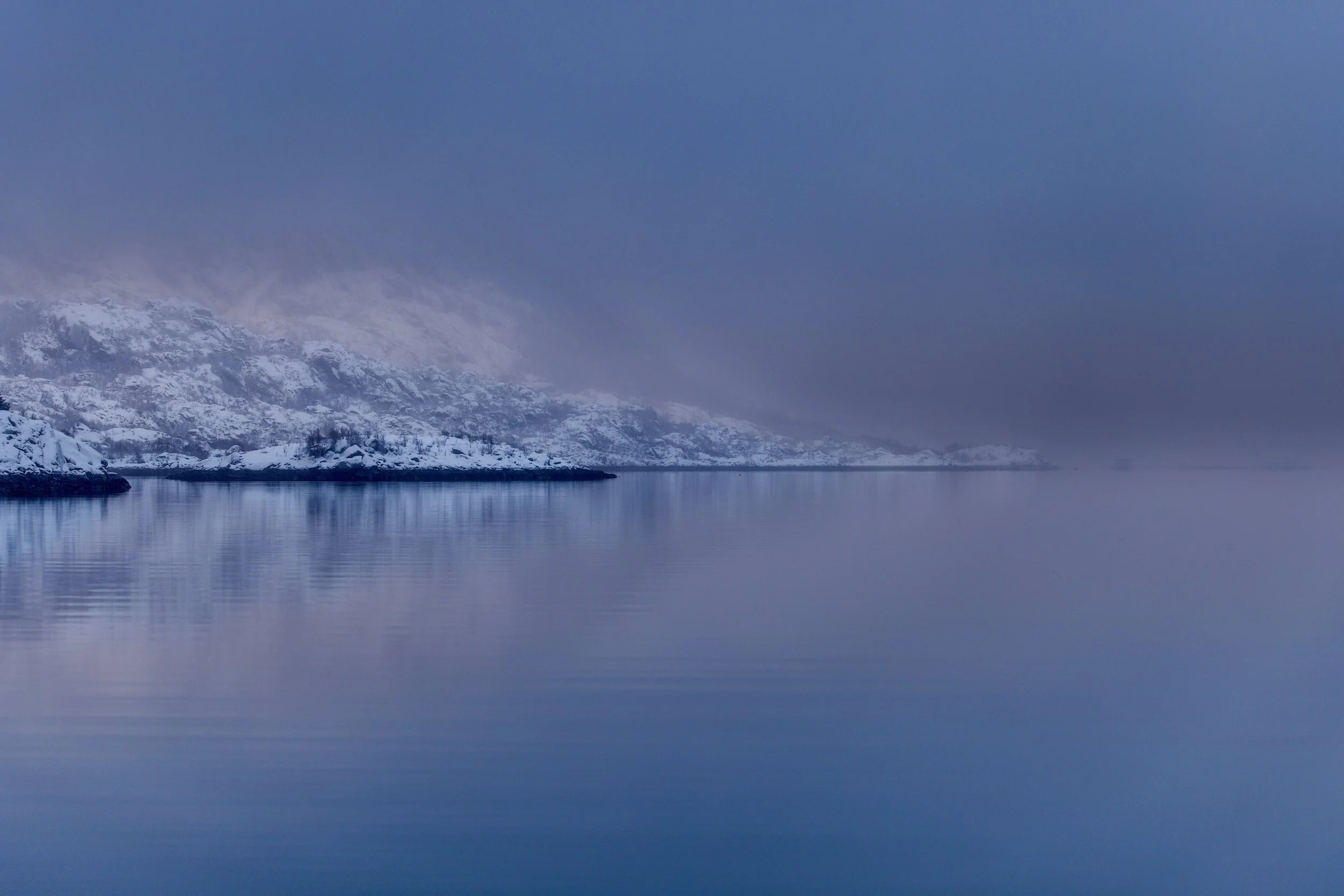 Snow-covered mountains and rocks along a calm, reflective body of water under a foggy, overcast sky.